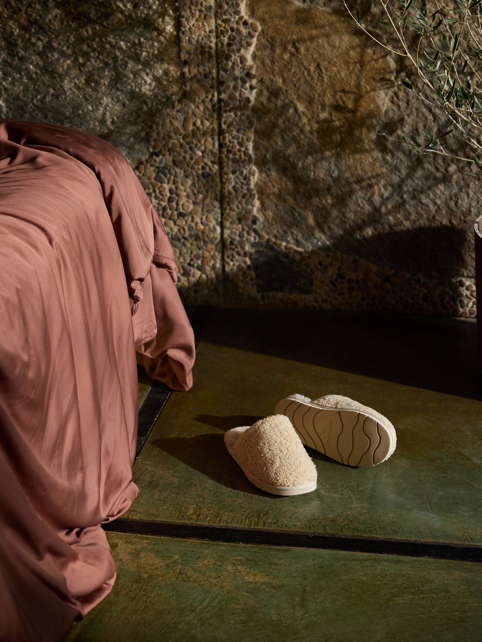 A pair of Cozy Earth Puffy Sheep Slippers are placed on a glossy, green floor next to a bed covered with a rust-colored, silky blanket. The background features a textured stone wall and part of a potted plant.