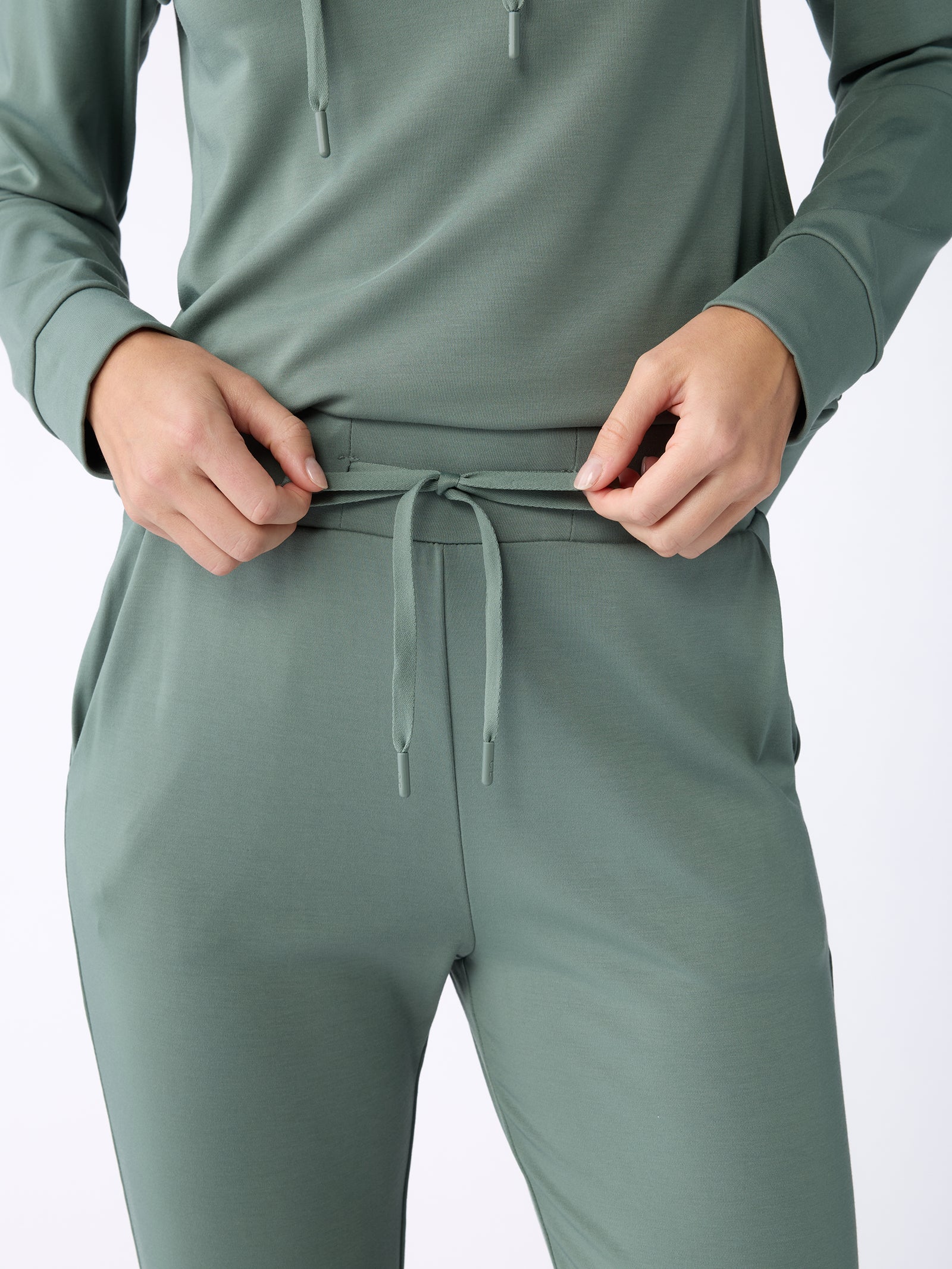 A person wearing the Women's Bamboo Jogger Pant TALL in green from Cozy Earth adjusts the drawstring on the pants. The image is focused on the torso and hands, highlighting the comfortable, casual outfit.
