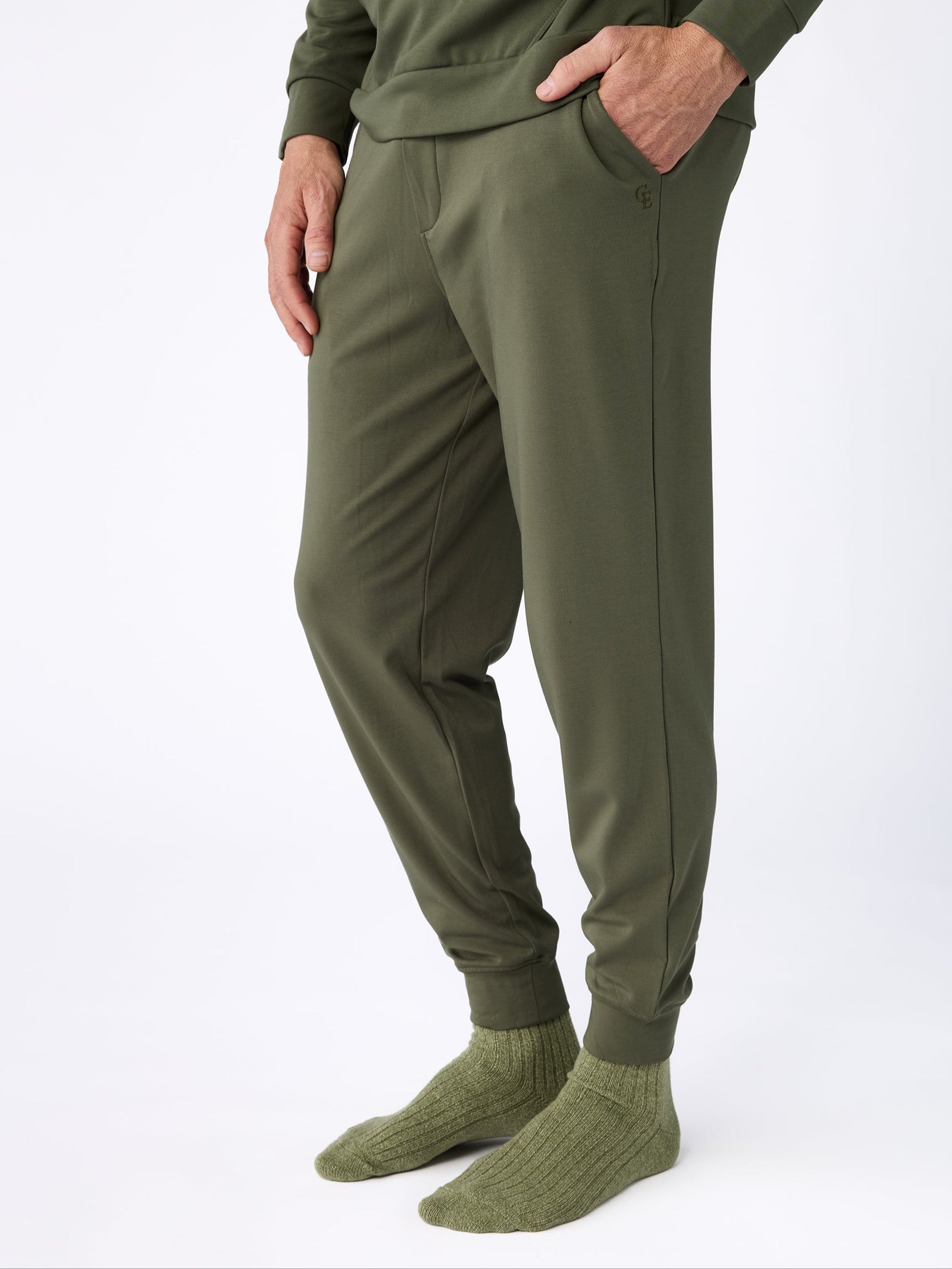 A person dressed completely in Cozy Earth's Men's Ultra-Soft Bamboo Pullover Crew in green stands against a white background. They have their hands in the pockets of their olive green pants and are wearing matching green socks.