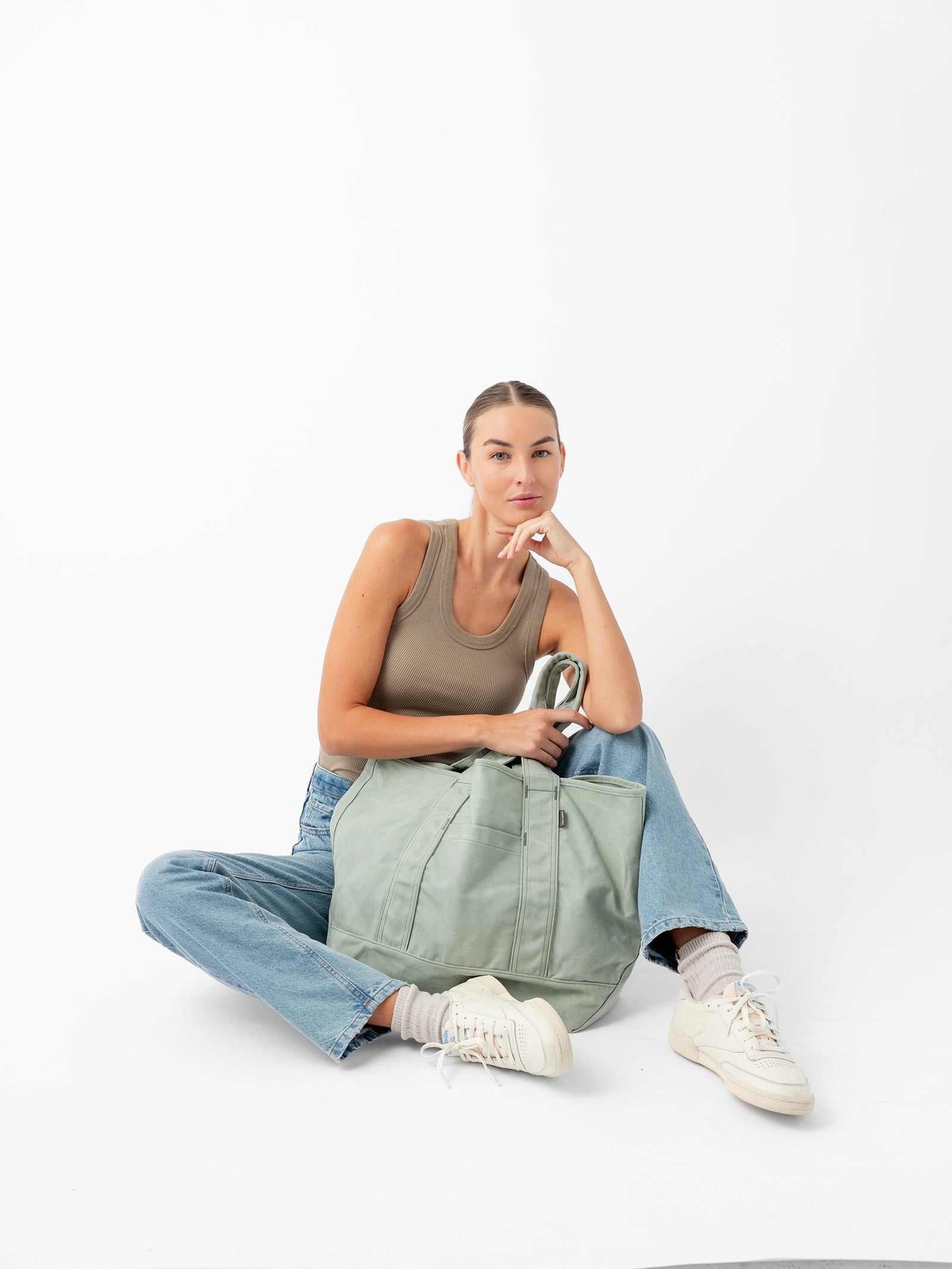 A woman with a pensive expression sits on the floor against a white background. She is wearing a beige tank top, blue jeans, and white sneakers. Resting her chin on her hand, she has a large Cozy Earth Waxed Canvas Tote in light green in front of her.