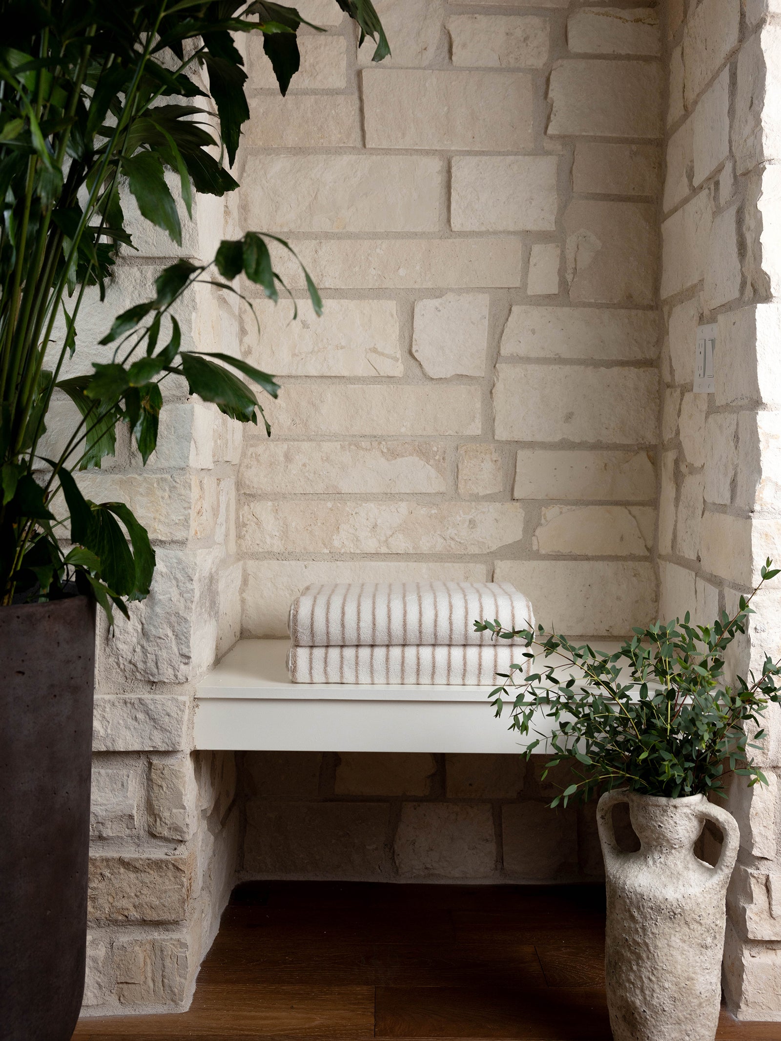 A cozy nook with a white stone wall features a minimalist cream-colored bench topped with two Luxe Bath Towels from Cozy Earth. The space is adorned with two lush green plants, one in a rustic gray pot and the other in an earthenware vase. Natural light highlights the serene setting.