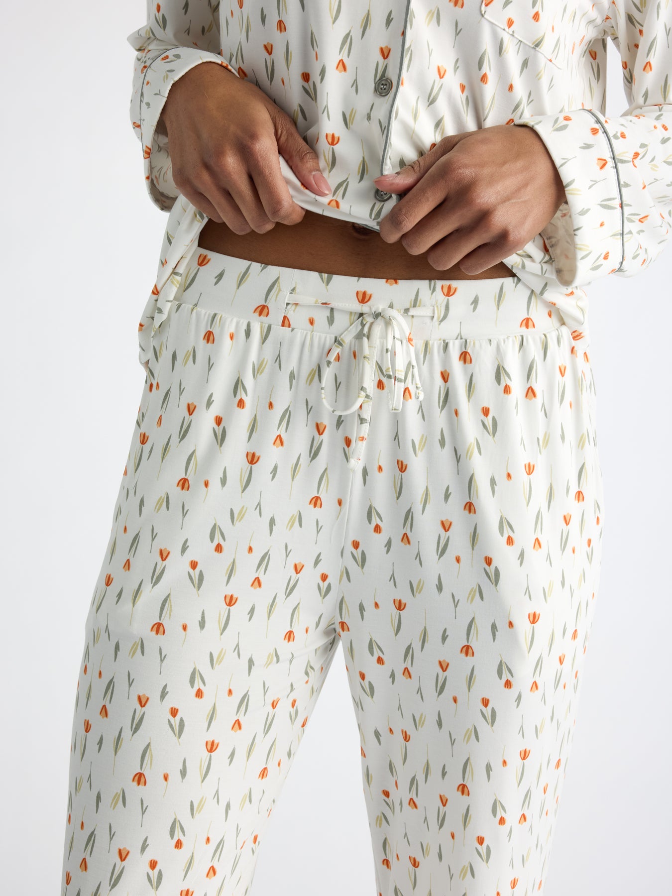 A person is wearing a matching pajama set featuring the Women's Bamboo Stretch-Knit Pajama Pant by Cozy Earth, showcasing a white background with small orange flowers. The focus is on their torso and hands, with one hand lifting the top slightly.
