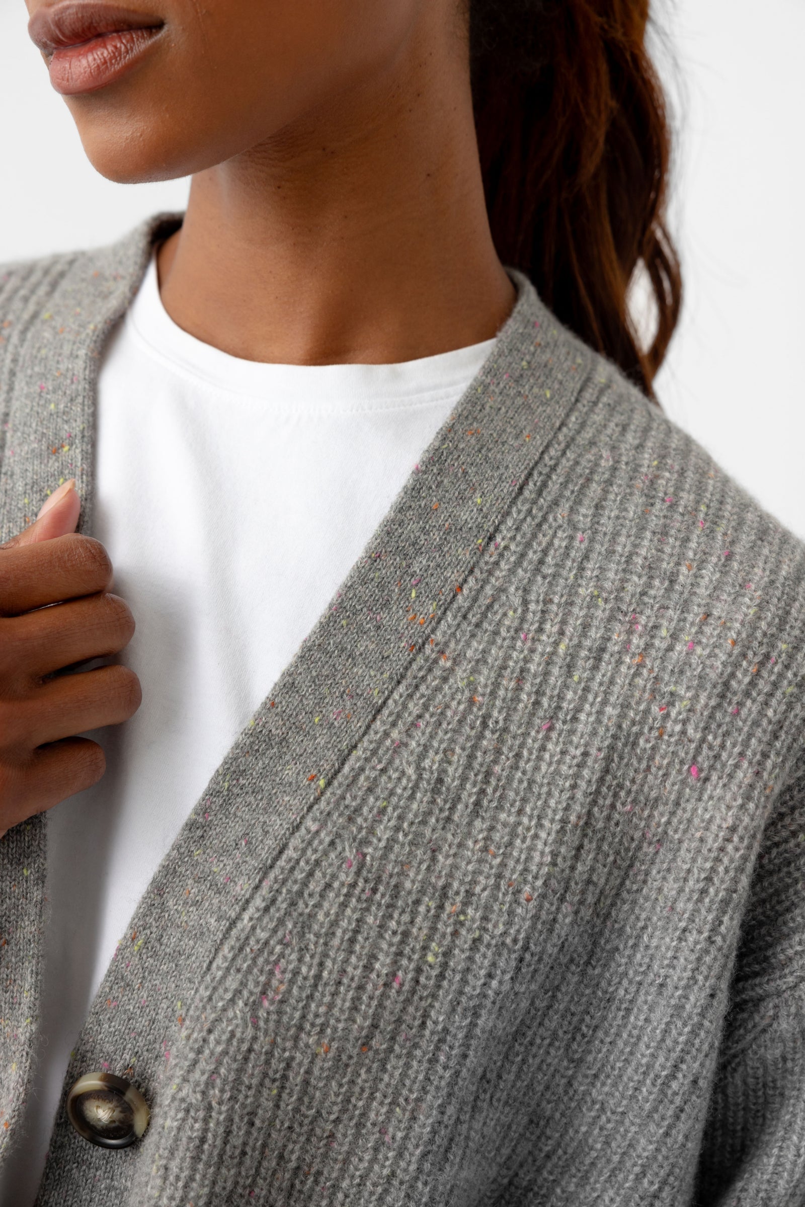 A person wearing the Oversized Sunday Cashmere Cardigan by Cozy Earth is partially visible, with their hand gently holding the cardigan closed over a white shirt. The cardigan displays a speckled pattern and a single visible button against a plain white background.