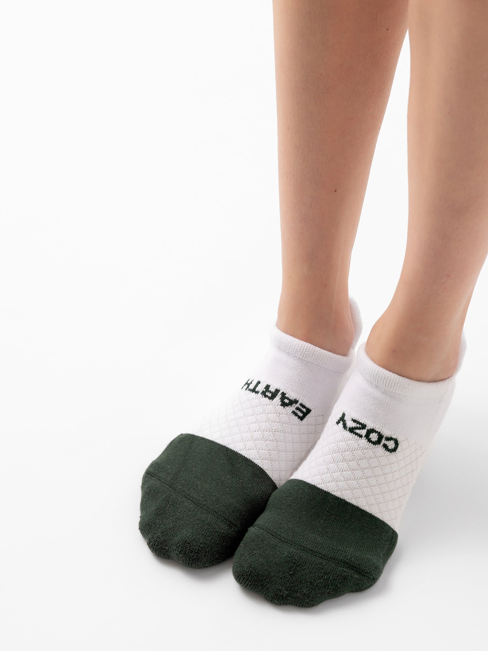 Close-up of a person's feet wearing white ankle socks from the "Essential Ankle Sock 2-Pack" by Cozy Earth. The socks feature dark green toes and heels, with "COZY EARTH" written on them—"COZY" on the right foot and "EARTH" on the left foot. The background is plain white.