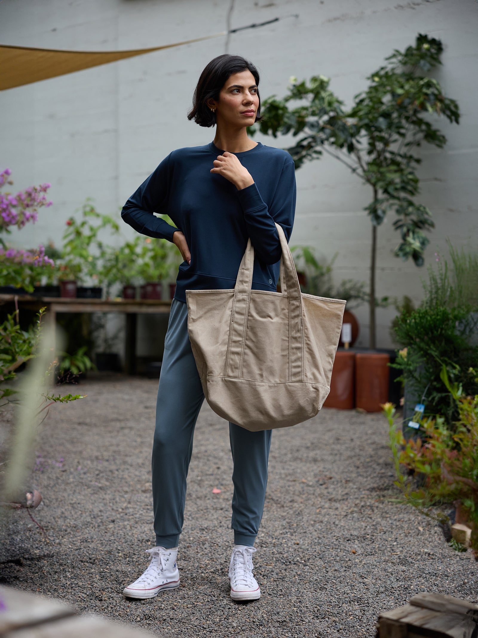 A person with short dark hair stands outdoors, carrying a large beige Waxed Canvas Tote from Cozy Earth over their shoulder. They wear a dark long-sleeve top, blue-gray pants, and white sneakers. Small potted plants and trees are in the background.