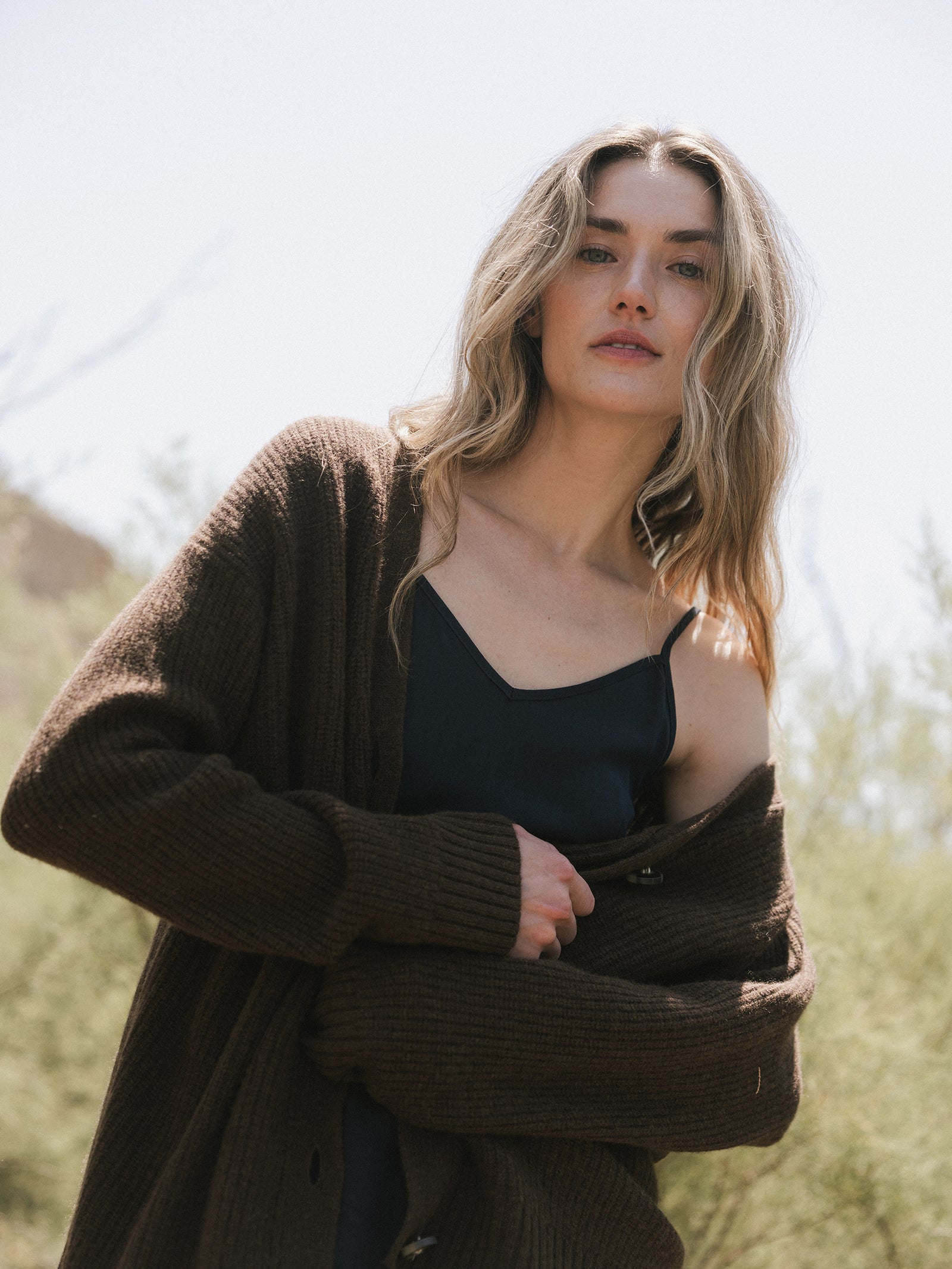 A person with long, wavy hair stands outdoors, wearing the Oversized Sunday Cashmere Cardigan from Cozy Earth over a black top. The background features soft-focus greenery and a clear sky, creating a serene and natural setting.