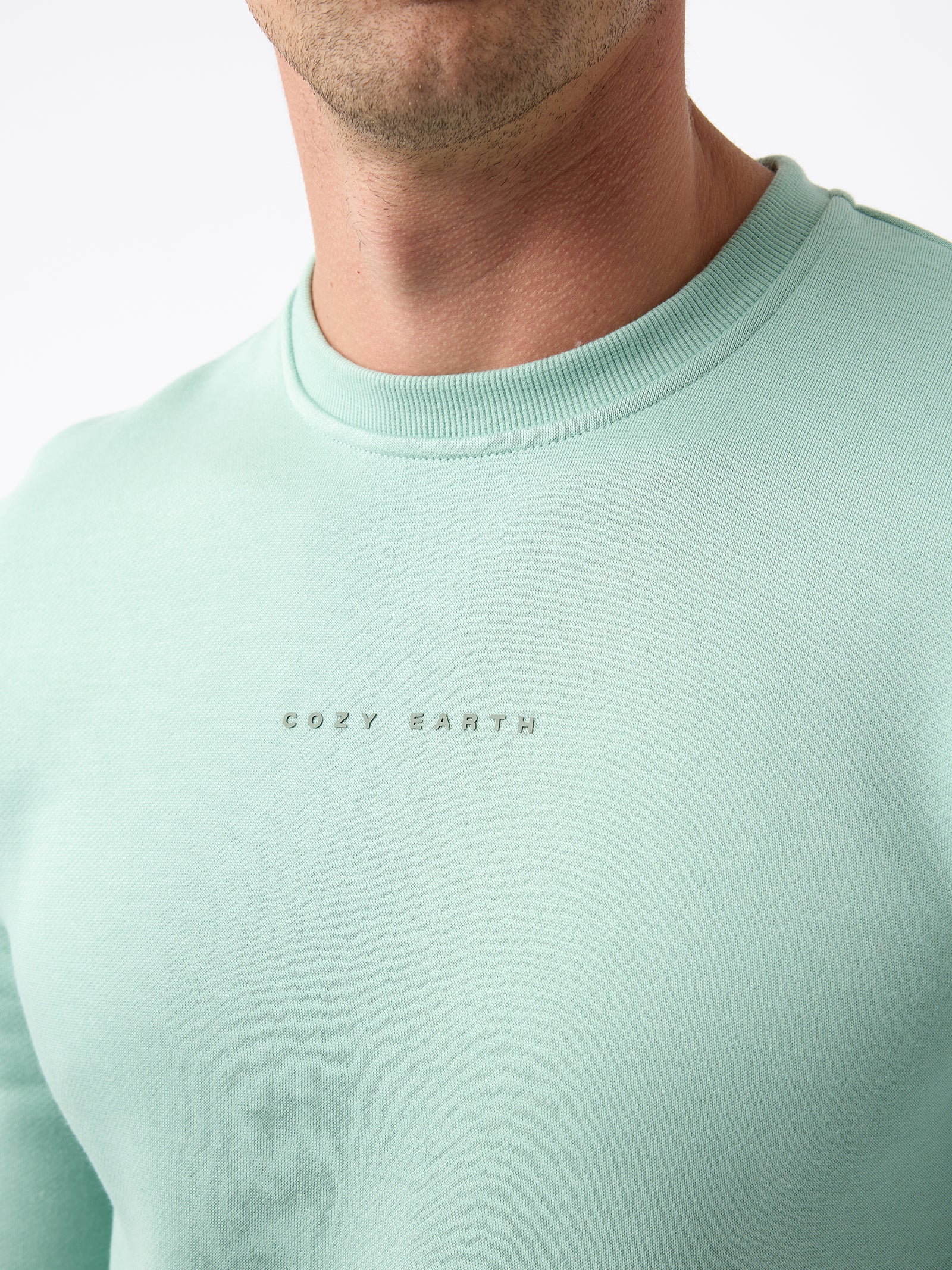 A person wearing a Men's CityScape Crewneck in light green with "Cozy Earth" printed on the front. The focus is on the upper section of the clothing, set against a plain white background.