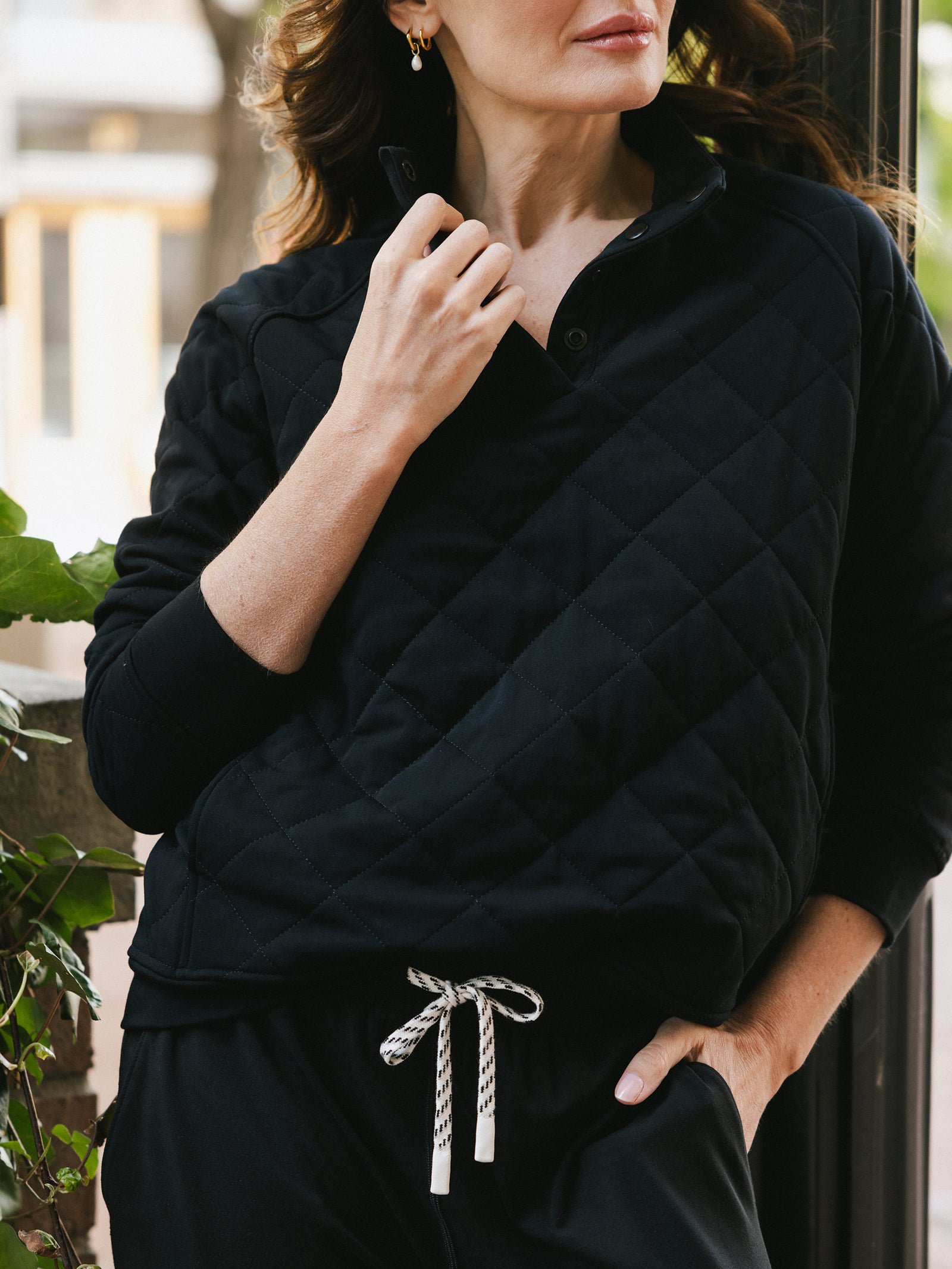 A person wearing a Cozy Earth Women's Ultra-Soft Bamboo Quilted Snap Pullover in black, along with matching black pants featuring a white and black drawstring, stands near a plant. The person is holding the pullover's collar with one hand while their other hand rests in their pocket. Their face is partially visible.
