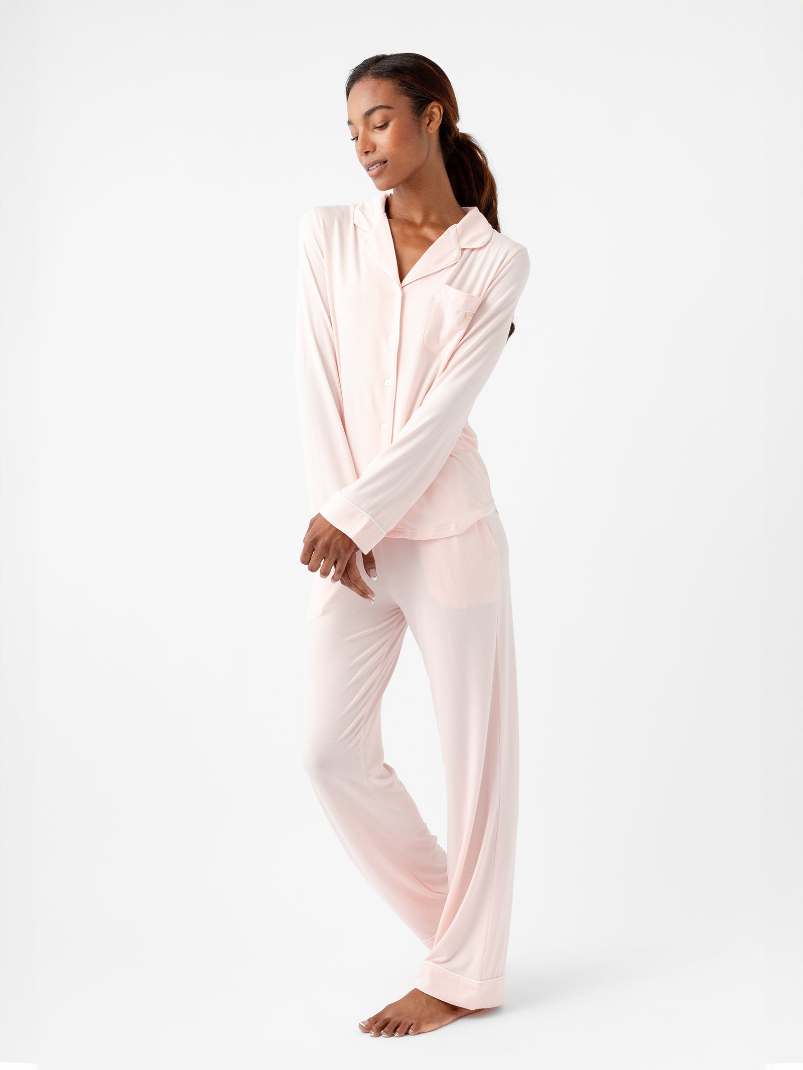 A woman stands barefoot against a plain white background, wearing the Women's Long Sleeve Bamboo Pajama Top in Stretch-Knit TALL from HIDE, in light pink. The pajama top has a collar and buttons, and the matching pants are loose-fitting. She has her arms folded slightly and looks down with a relaxed expression.
