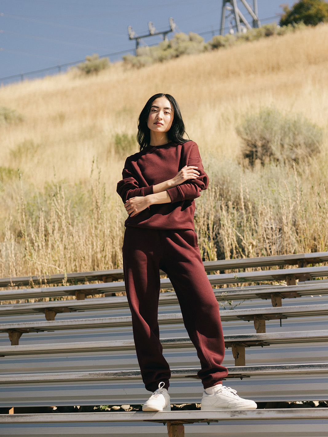 A person stands outdoors on metal bleachers, wearing a maroon Women's CityScape Sweatpant and matching sweatshirt by Cozy Earth. The background features dry grass and shrubs with power lines visible in the distance. The person has long dark hair and is looking confidently into the camera.