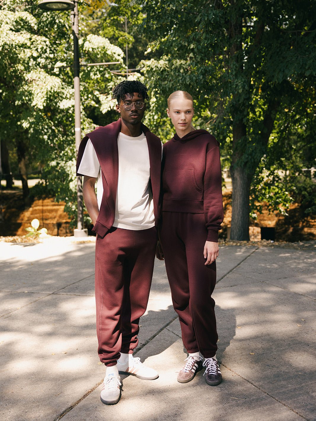 Two individuals stand in an outdoor setting with trees in the background, both wearing Cozy Earth's Men's CityScape Sweatpant in burgundy. The person on the left has a white t-shirt underneath and white sneakers, while the individual on the right sports matching burgundy sneakers. Both look at the camera.