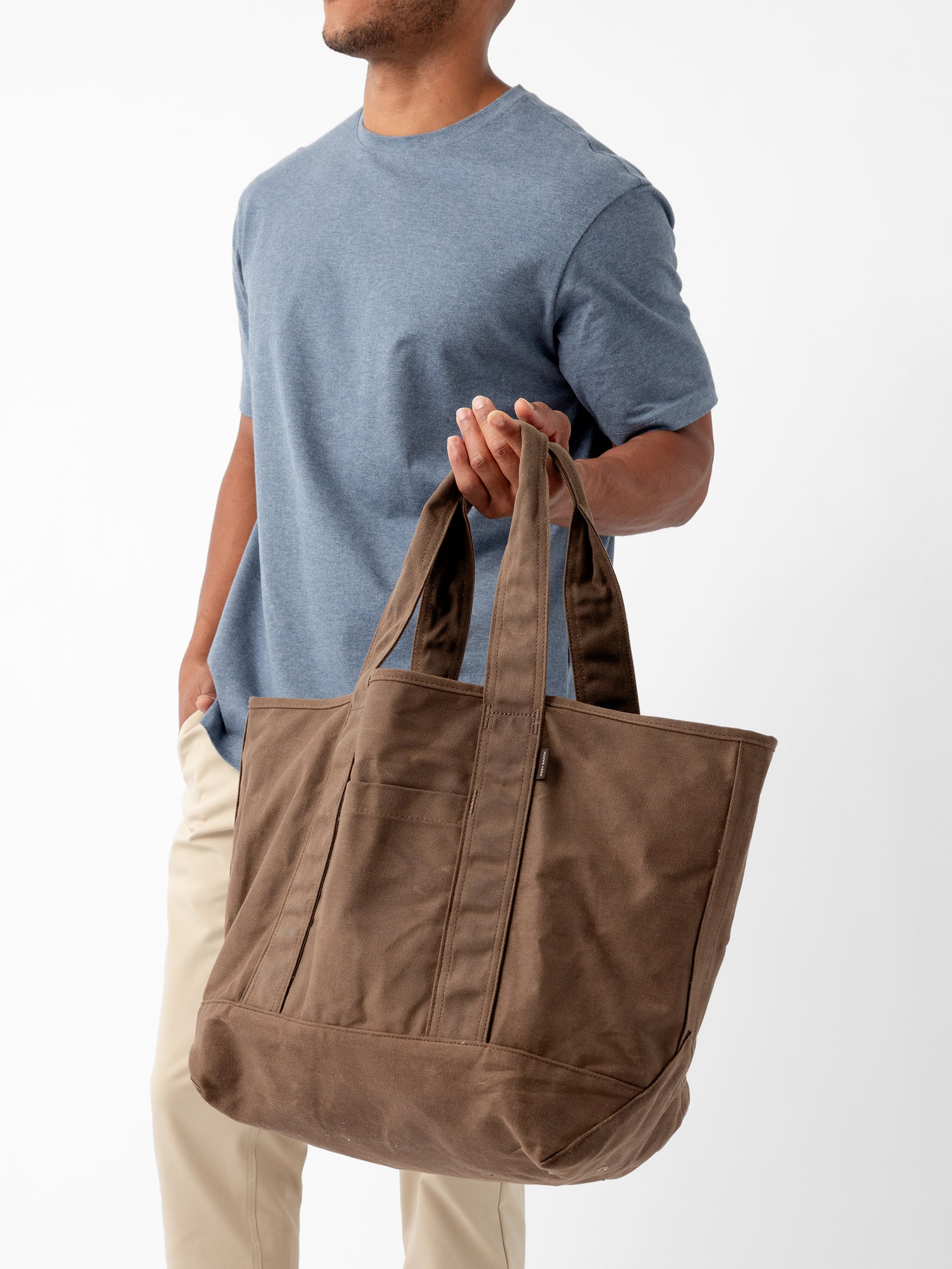 A person in a blue t-shirt and beige pants holds a large Waxed Canvas Tote by Cozy Earth. The tote bag has long straps and appears spacious. The background is plain white. The person's face is not visible.