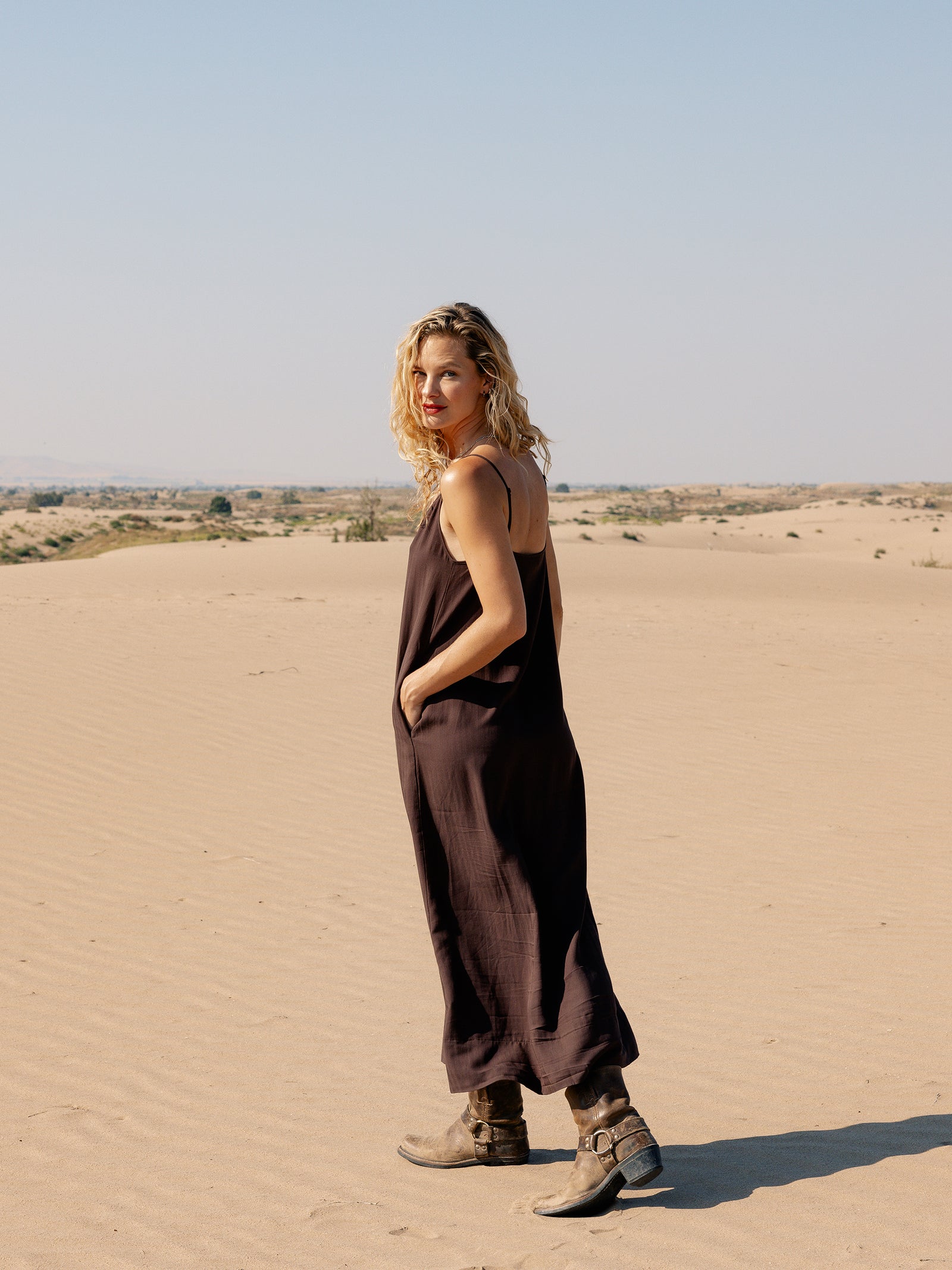A woman with curly blonde hair, wearing a loose Women’s Sunset Slip Dress from Cozy Earth and brown boots, stands in a sandy desert landscape. She looks over her shoulder at the camera with a slight smile. The sky is clear and blue.