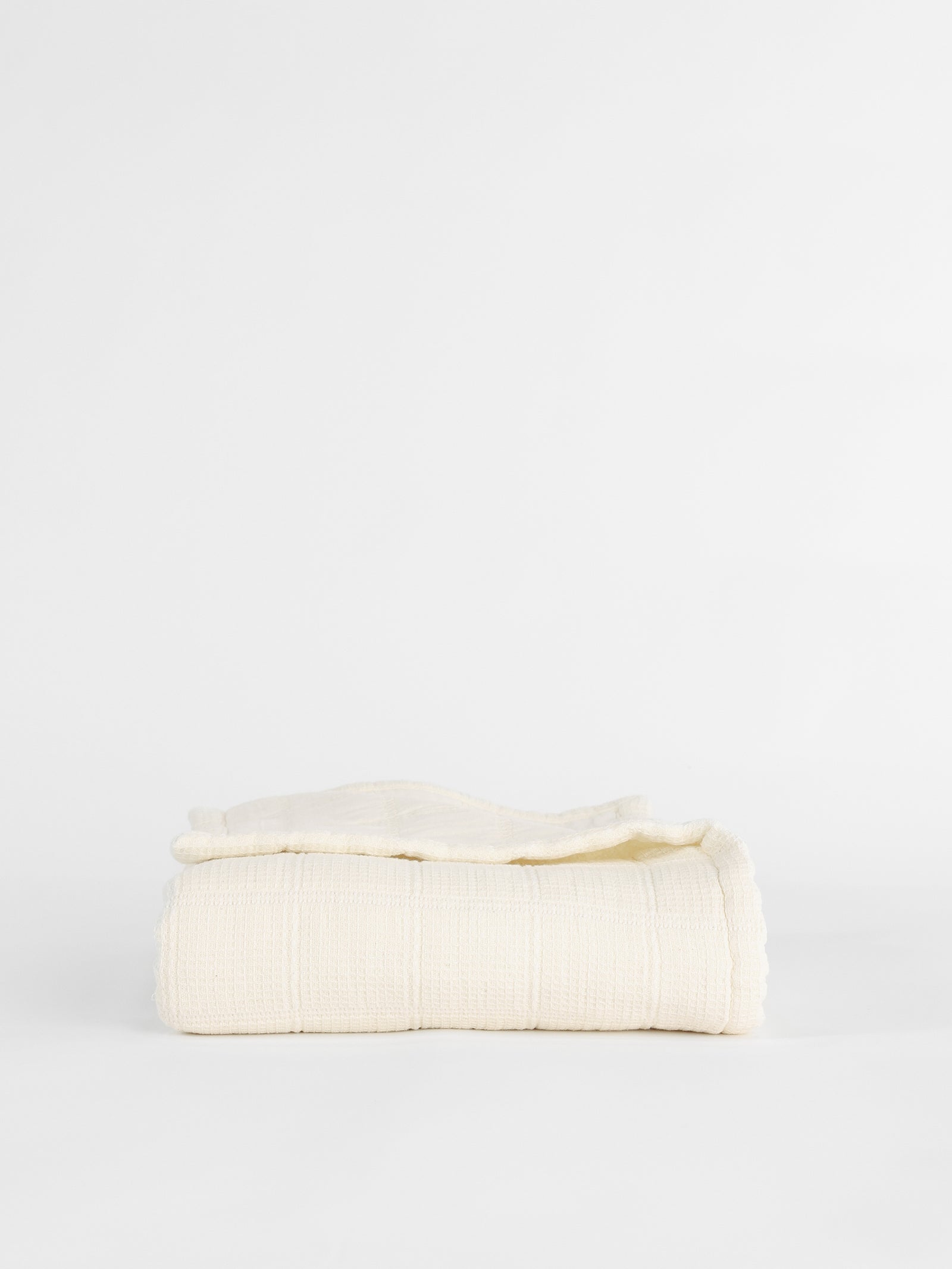 A neatly folded Cozy Earth Waffle Windowpane Blanket, available in beige or off-white and featuring a textured grid-like pattern, is placed against a plain white background.
