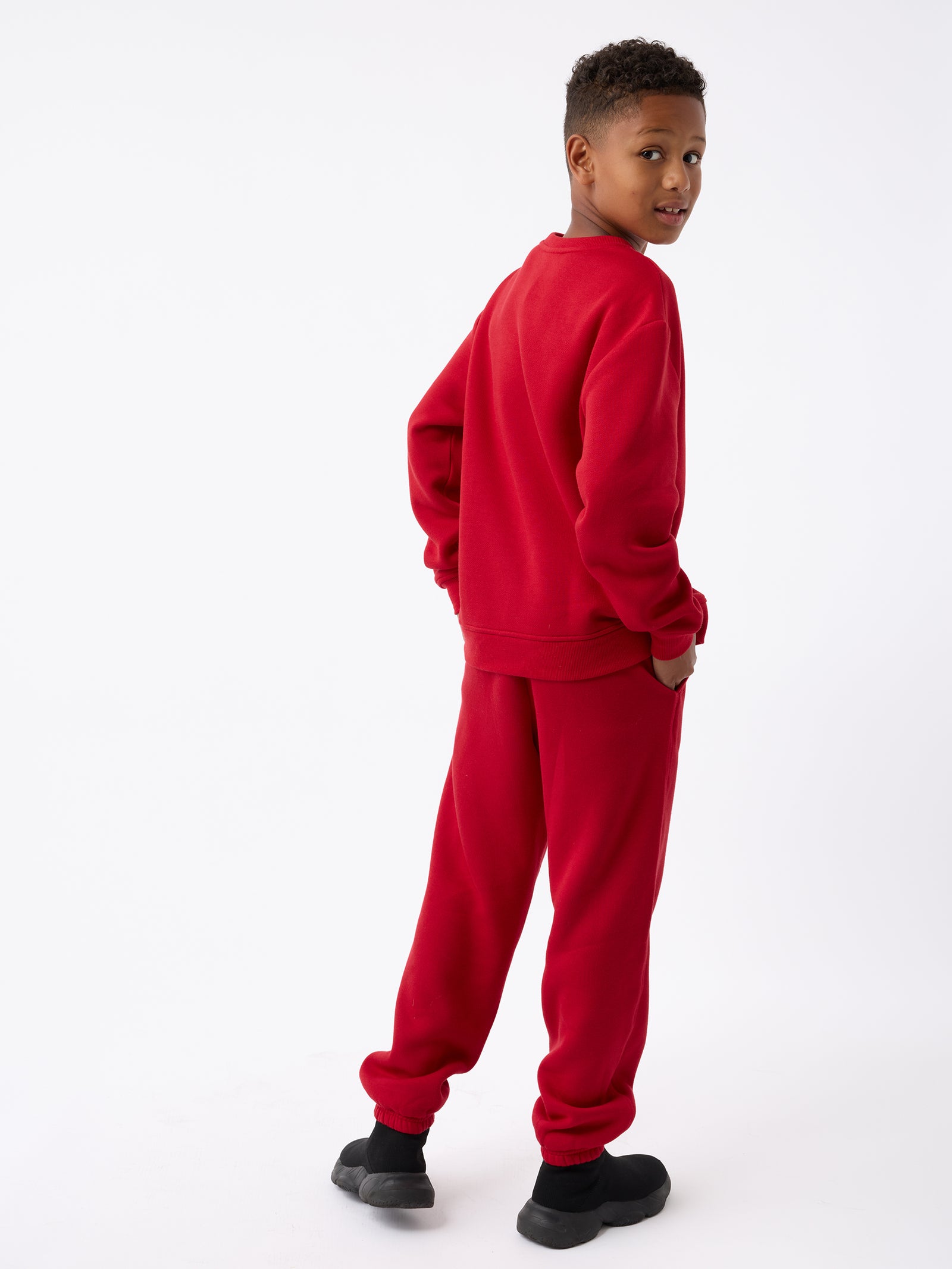 A young boy wearing the Kid's CityScape Crewneck by Cozy Earth and black shoes stands with his back turned, looking over his shoulder and smiling against a plain white background.