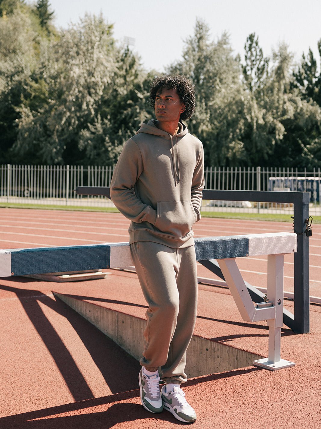 A person wearing a Cozy Earth Men's CityScape Sweatpant and a matching tan hoodie leans against a hurdle on an outdoor track, gazing away from the camera. Trees and a metal fence are visible in the background beneath a clear sky.