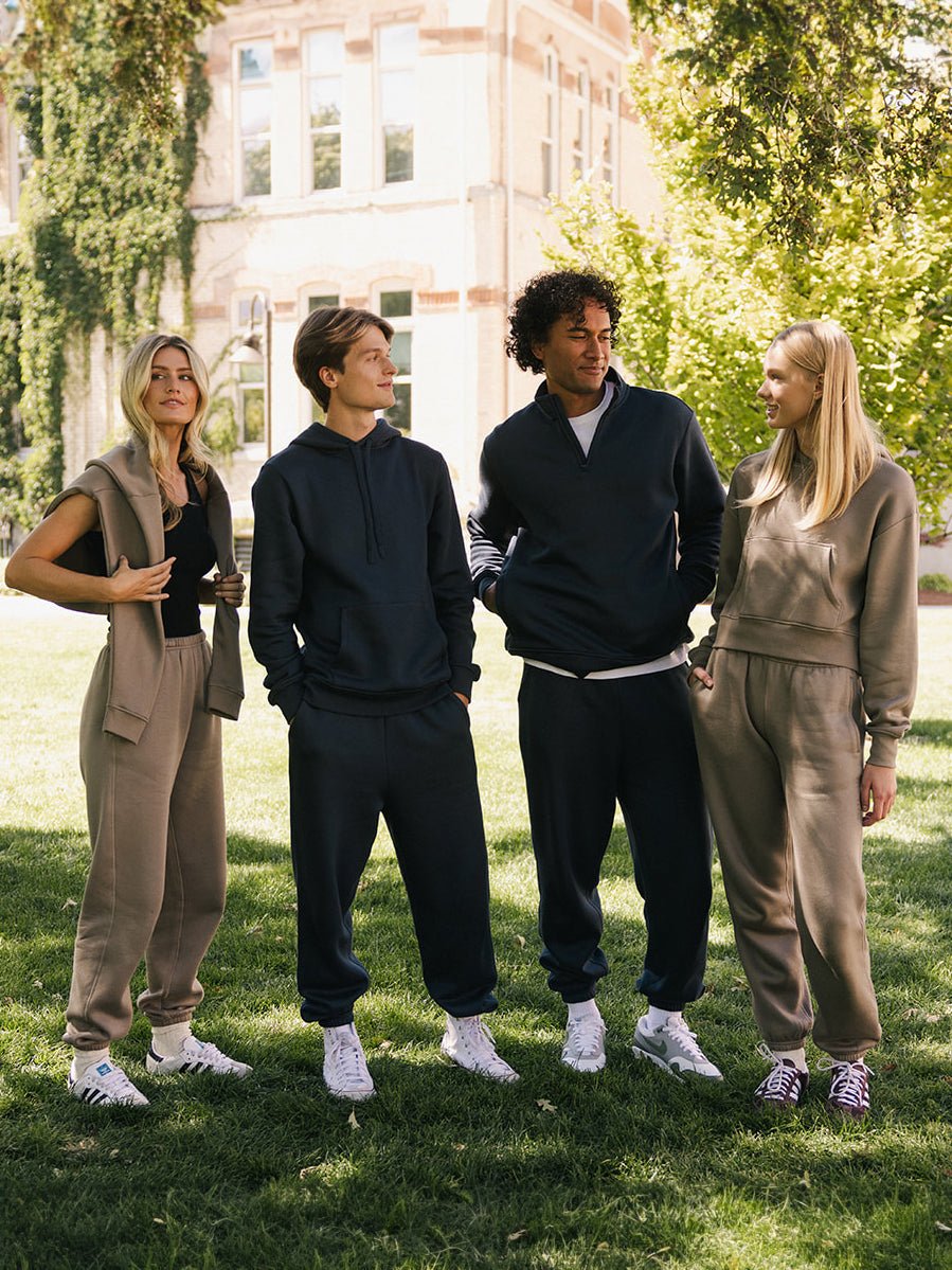 Four people stand conversing on a grassy lawn with trees and a building in the background. They are wearing casual, matching outfits consisting of Cozy Earth's Men's CityScape Sweatpant in earthy tones and white sneakers. The weather appears sunny and pleasant.