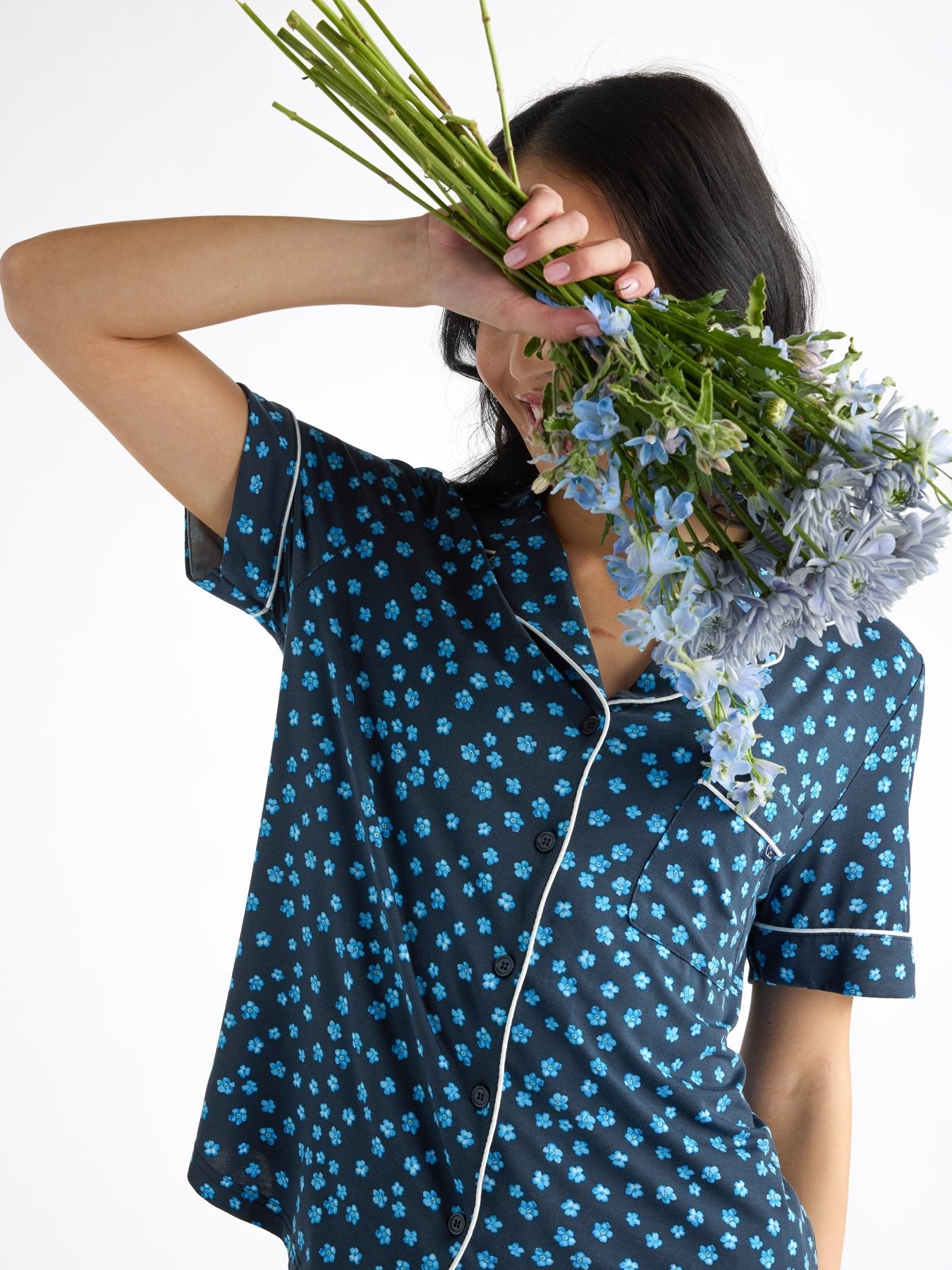 A person in a blue floral-patterned women's bamboo stretch-knit short sleeve pajama set by Cozy Earth playfully holds a bouquet of blue flowers, partially covering their face, with loose dark hair and a plain white background.