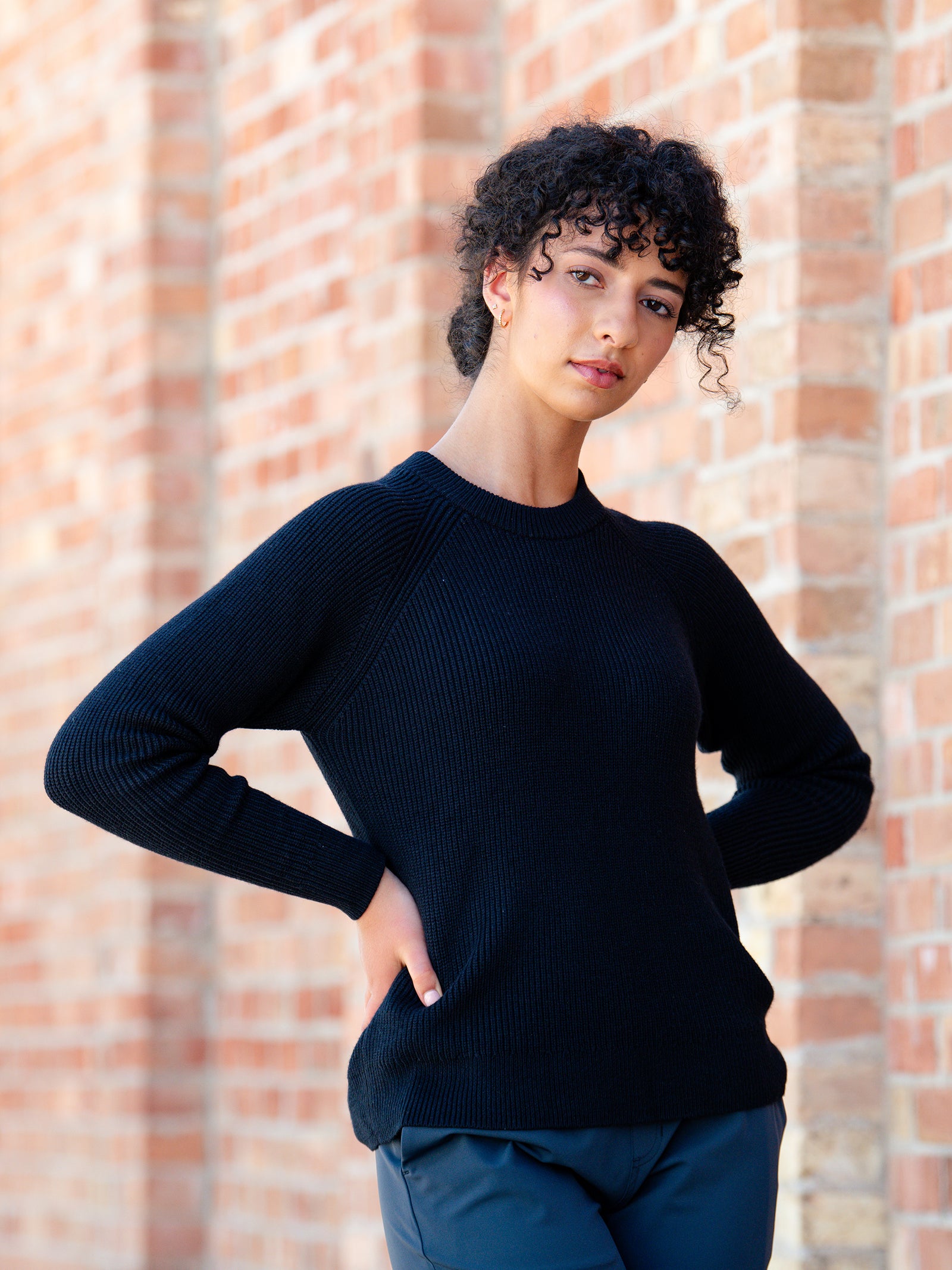 A person with curly hair stands against a brick wall, posing with one hand on their hip and the other by their side. They are wearing a Cozy Earth Women's Classic Crewneck and dark pants. The expression on their face is neutral.