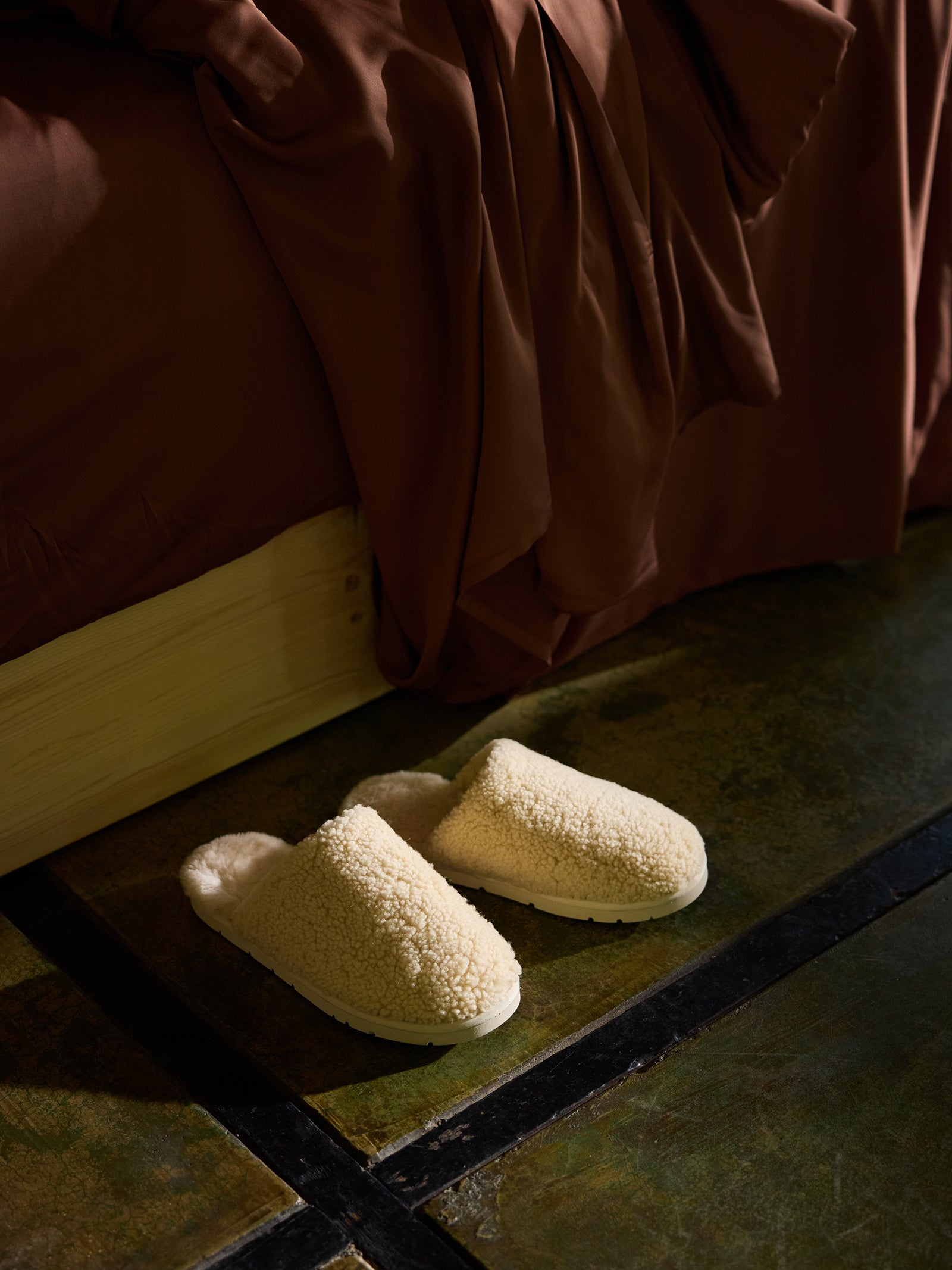 A pair of Cozy Earth's Puffy Sheep Slippers, in a cozy, cream-colored, fluffy fabric, lie on a mottled green floor, partially shaded. They are positioned next to a tan wooden bed frame with dark brown bedding draped over the side. The scene appears warm and inviting, suggestive of a cozy home environment.
