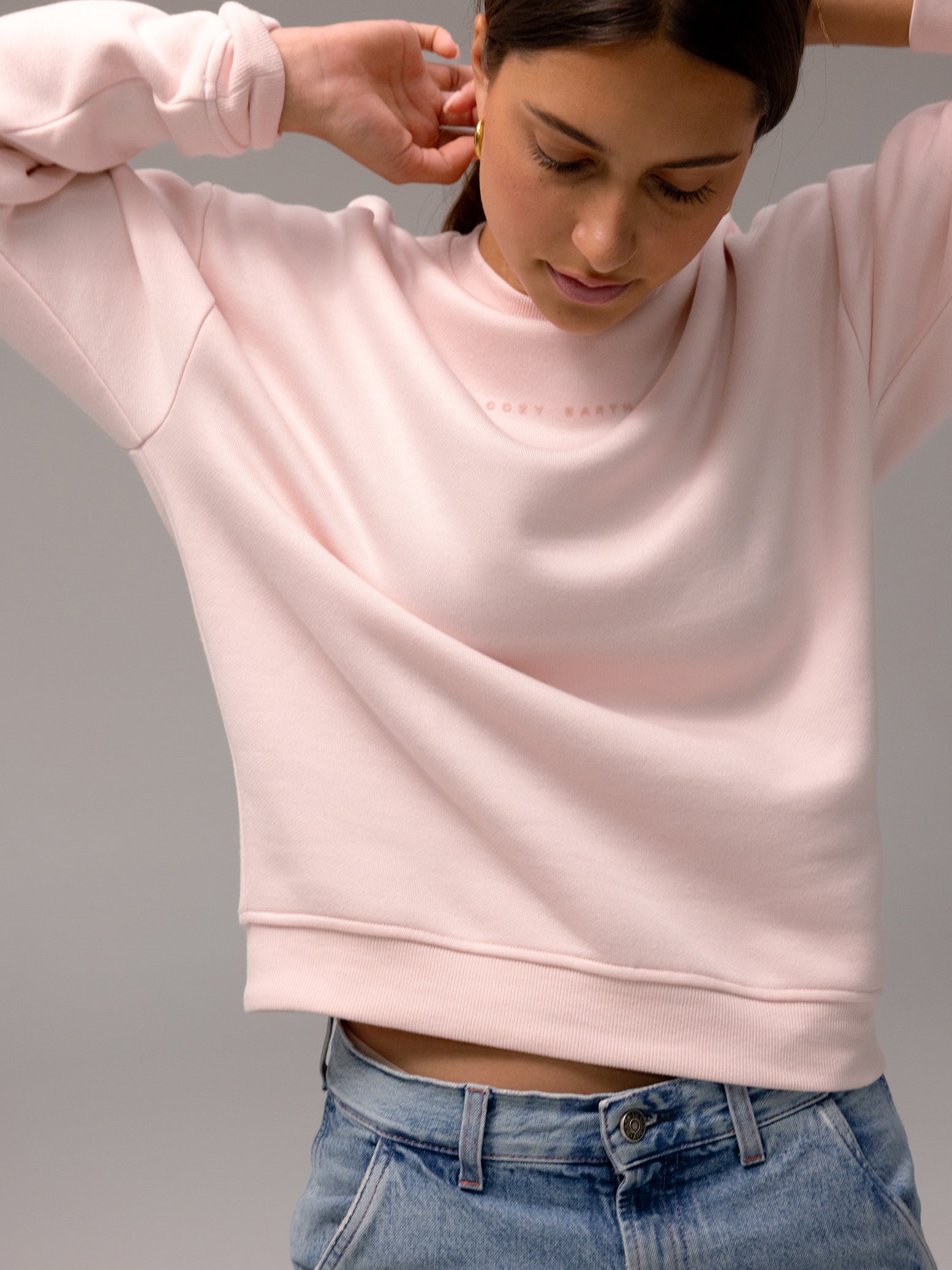 A woman wearing the light pink Women's CityScape Crewneck by Cozy Earth and blue jeans leans forward, looking down, against a plain gray background.