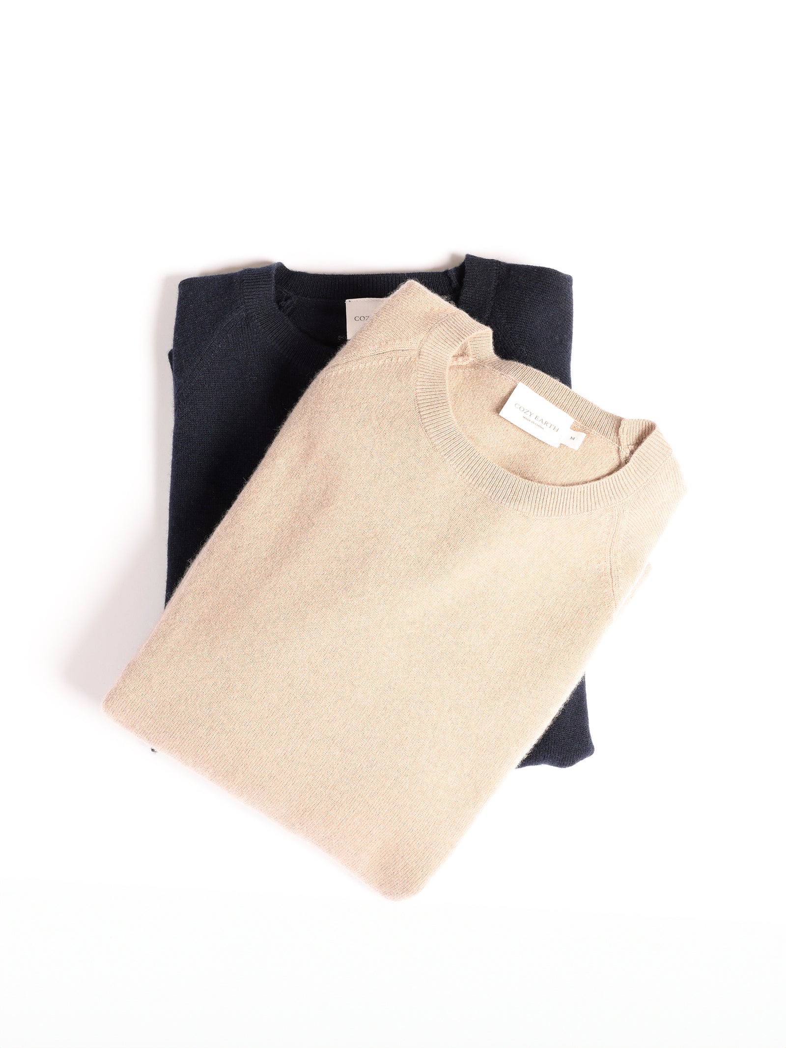 Two neatly folded Cozy Earth Men's Crewneck Sweaters placed on a white background. The top sweater is light beige, and the one underneath is dark navy. Each sweater has a small white label near the collar with text on it.