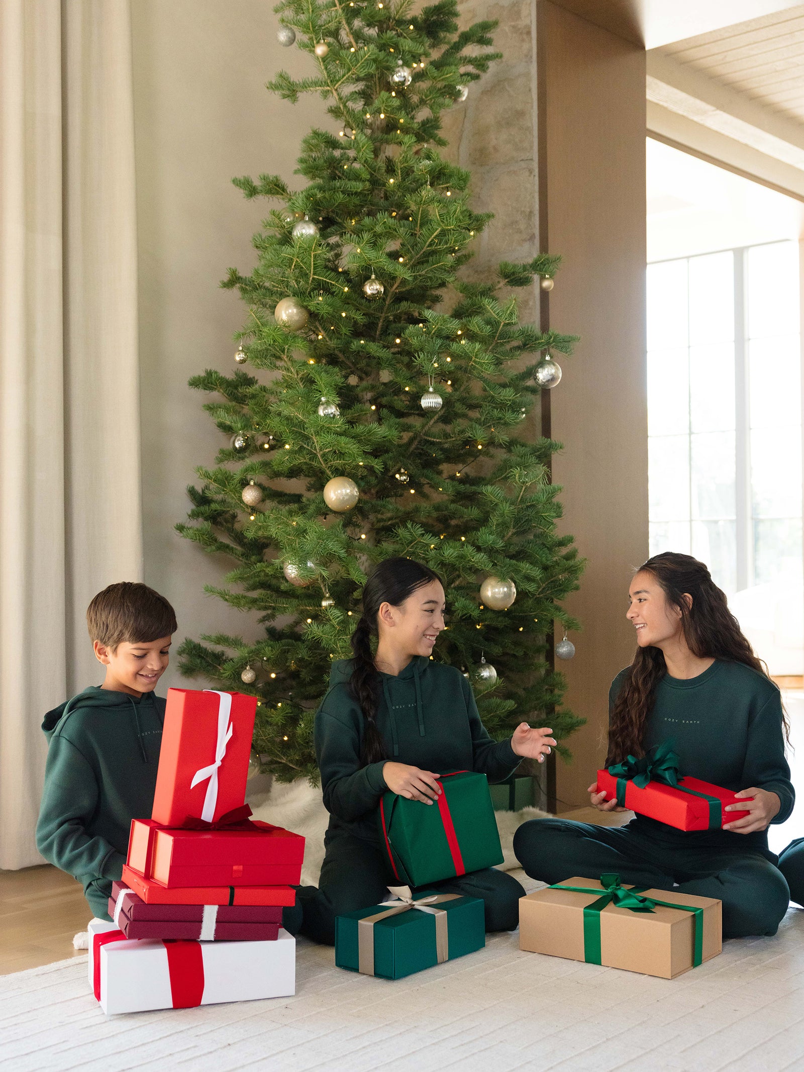 A family of three sits by a decorated Christmas tree, engaging in gift-wrapping. They smile while surrounded by colorful presents, including Cozy Earth's Kid's CityScape Crewneck. The room is bright and cozy, enhancing the festive atmosphere.