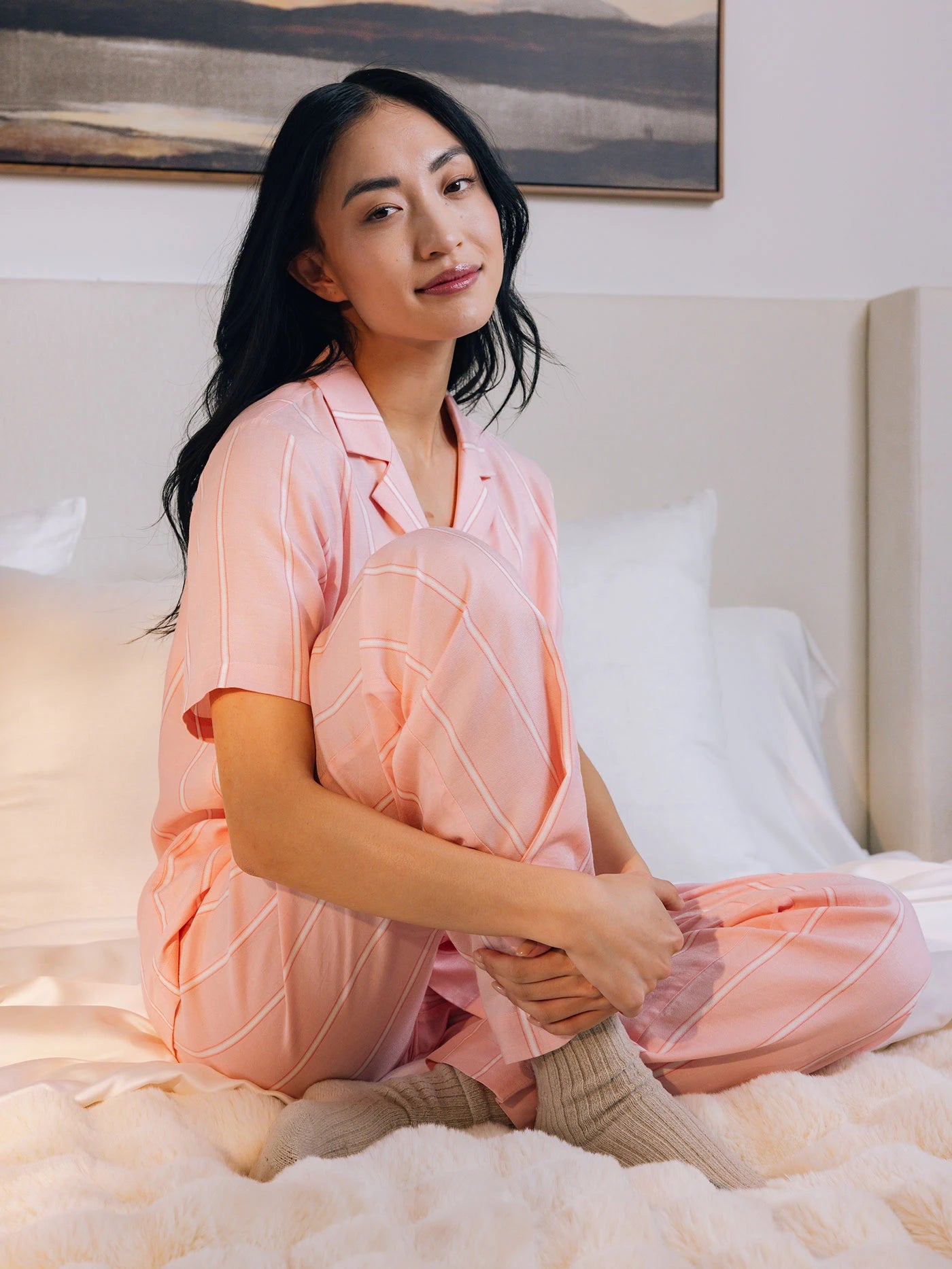 A person with long dark hair sits cross-legged on a bed wearing Cozy Earth's Women's Soft Woven Pajama Pant in pink and white stripes and wool socks, gazing at the camera. The softly lit room features a framed picture in the background.
