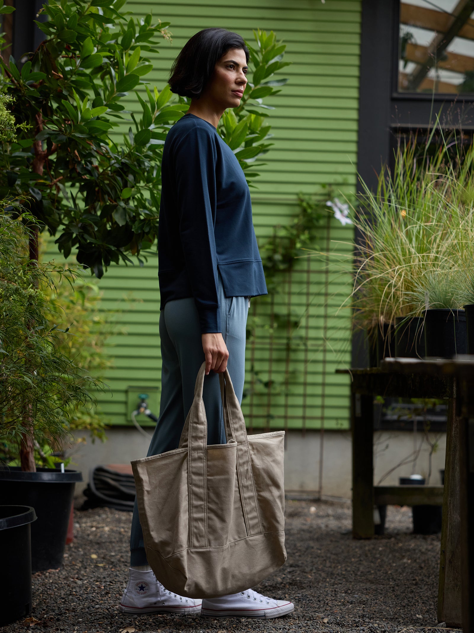 A person in a navy sweater and light blue pants stands in a garden area holding a large Waxed Canvas Tote by Cozy Earth. They have short dark hair and are wearing white sneakers. The background features green plants, trees, and a green building with horizontal siding.