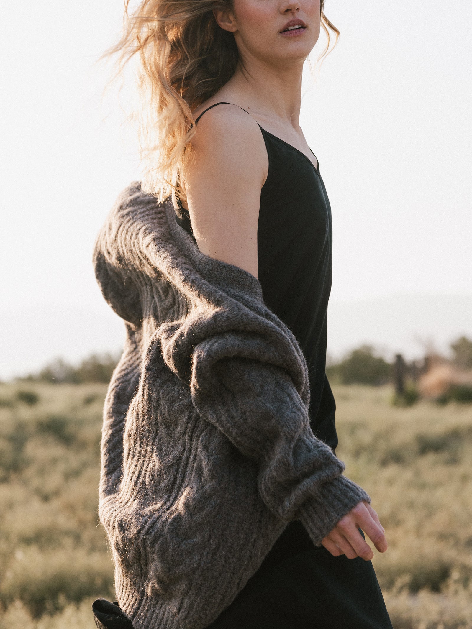 A person with wavy hair walks through a field, wearing a black dress and an Oversized Cable Knit Cardigan by Cozy Earth draped over their shoulder. The scene is serene with a soft, warm light.