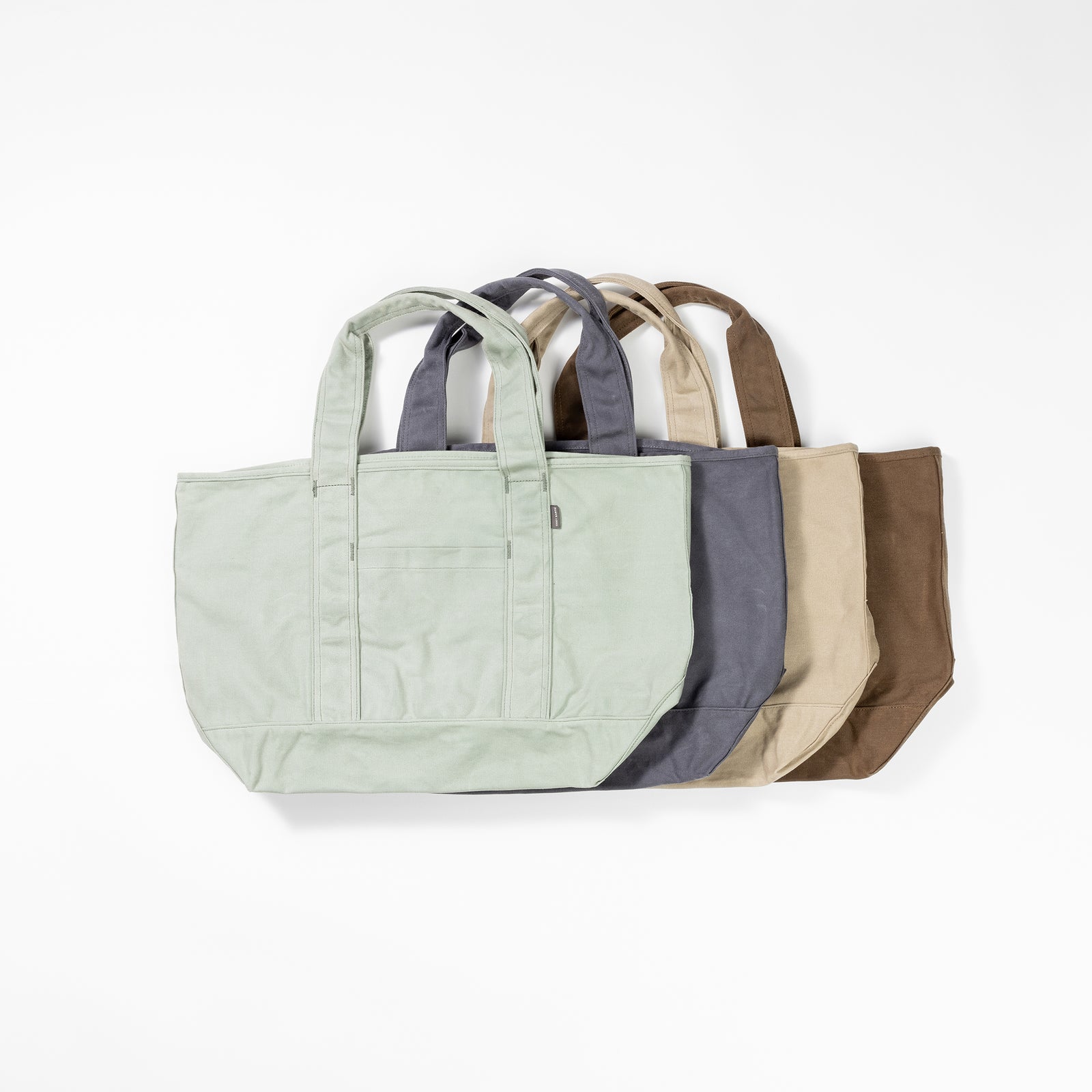 Five Waxed Canvas Totes from Cozy Earth, in light green, gray, beige, brown, and dark gray, are stacked side by side. Each tote has two handles and a front pocket against a plain white background.