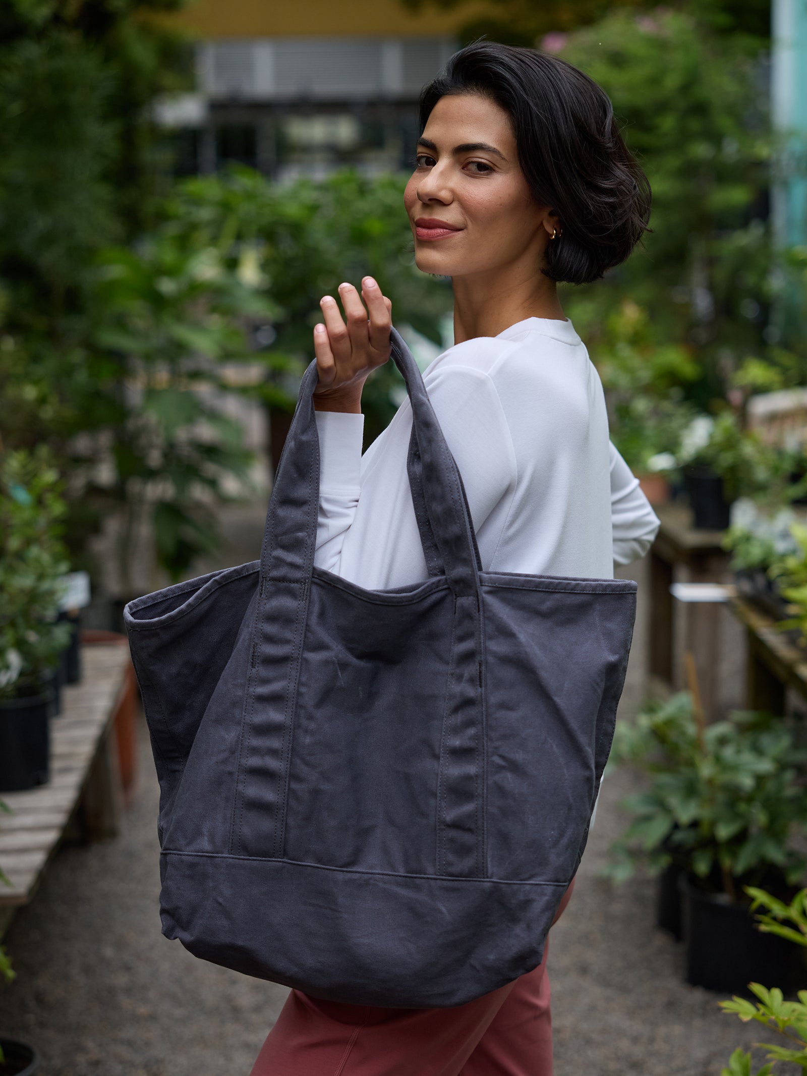 A woman with short dark hair, wearing a white long-sleeve shirt and pink pants, is standing in a lush garden center. She is smiling and looking over her shoulder while holding a large Waxed Canvas Tote from Cozy Earth. Various potted plants and greenery surround her.