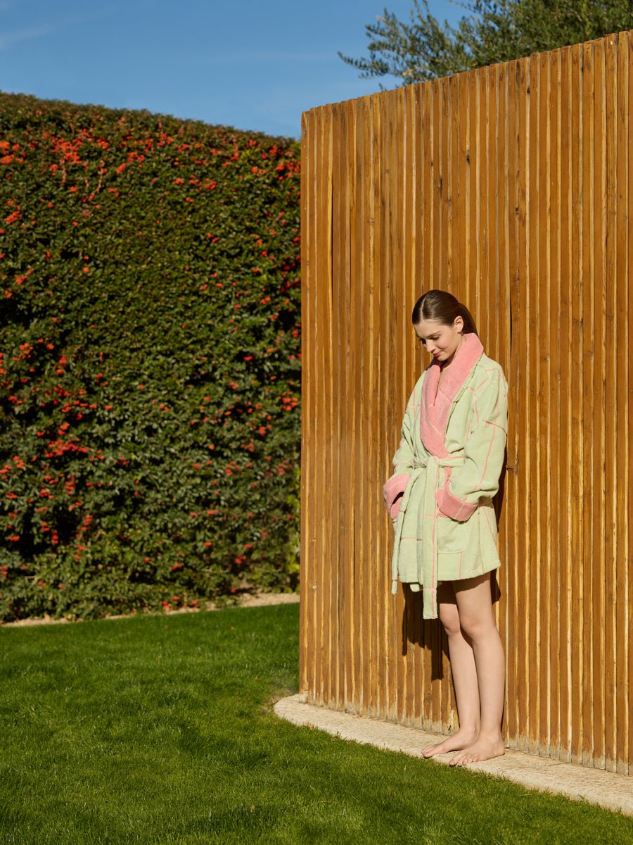 A woman wearing the Cozy Earth Windowpane Resort Robe in green with pink trim stands barefoot on grass, leaning against a wooden fence. A tall hedge with red flowers decorates the background beneath a clear blue sky.