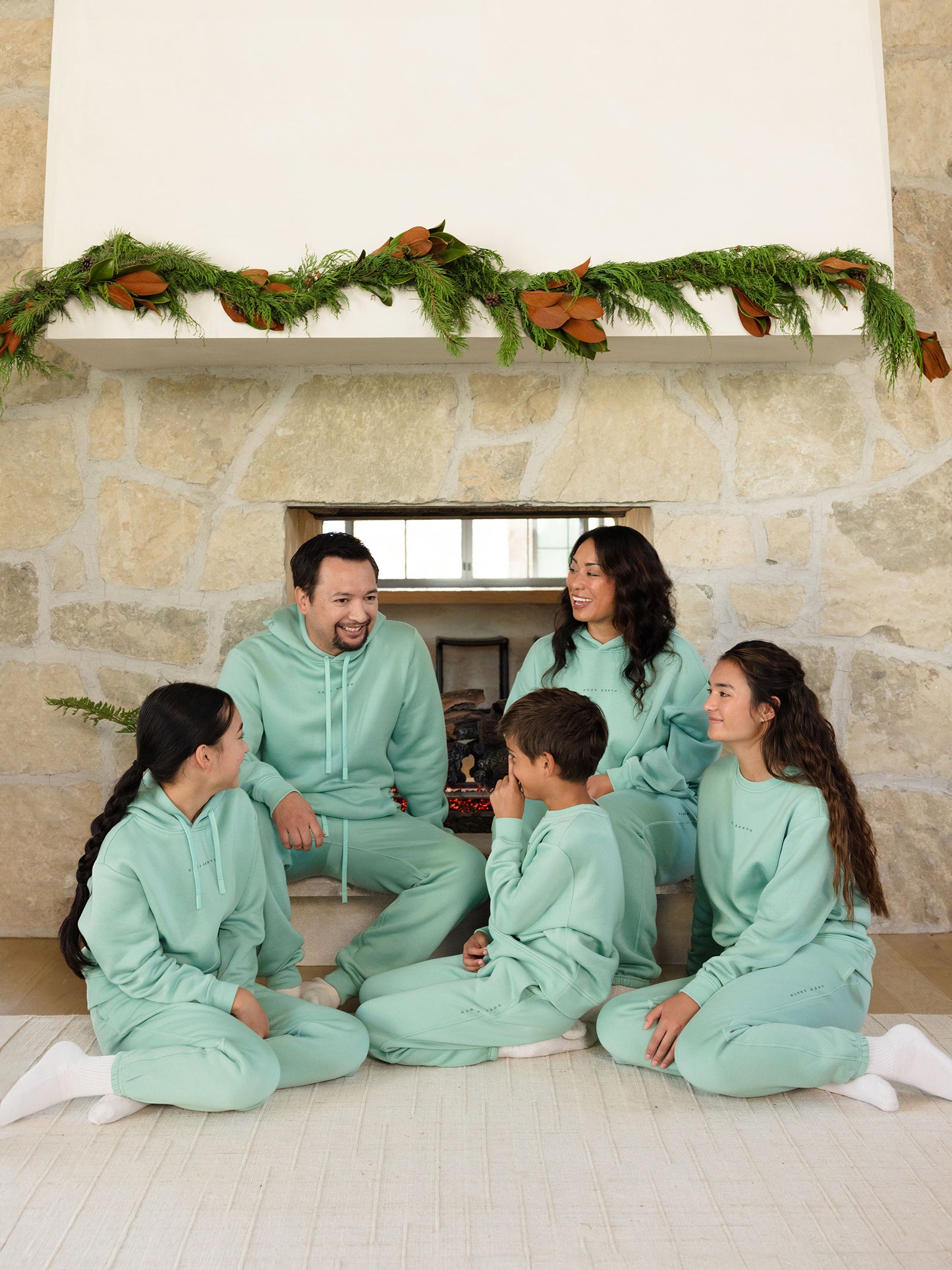 A family of five, dressed in matching mint green outfits including the Women's CityScape Sweatpant from Cozy Earth, sits in front of a fireplace adorned with a garland. They are sitting on a light rug, smiling and looking at each other. The room has stone walls and a warm ambiance.