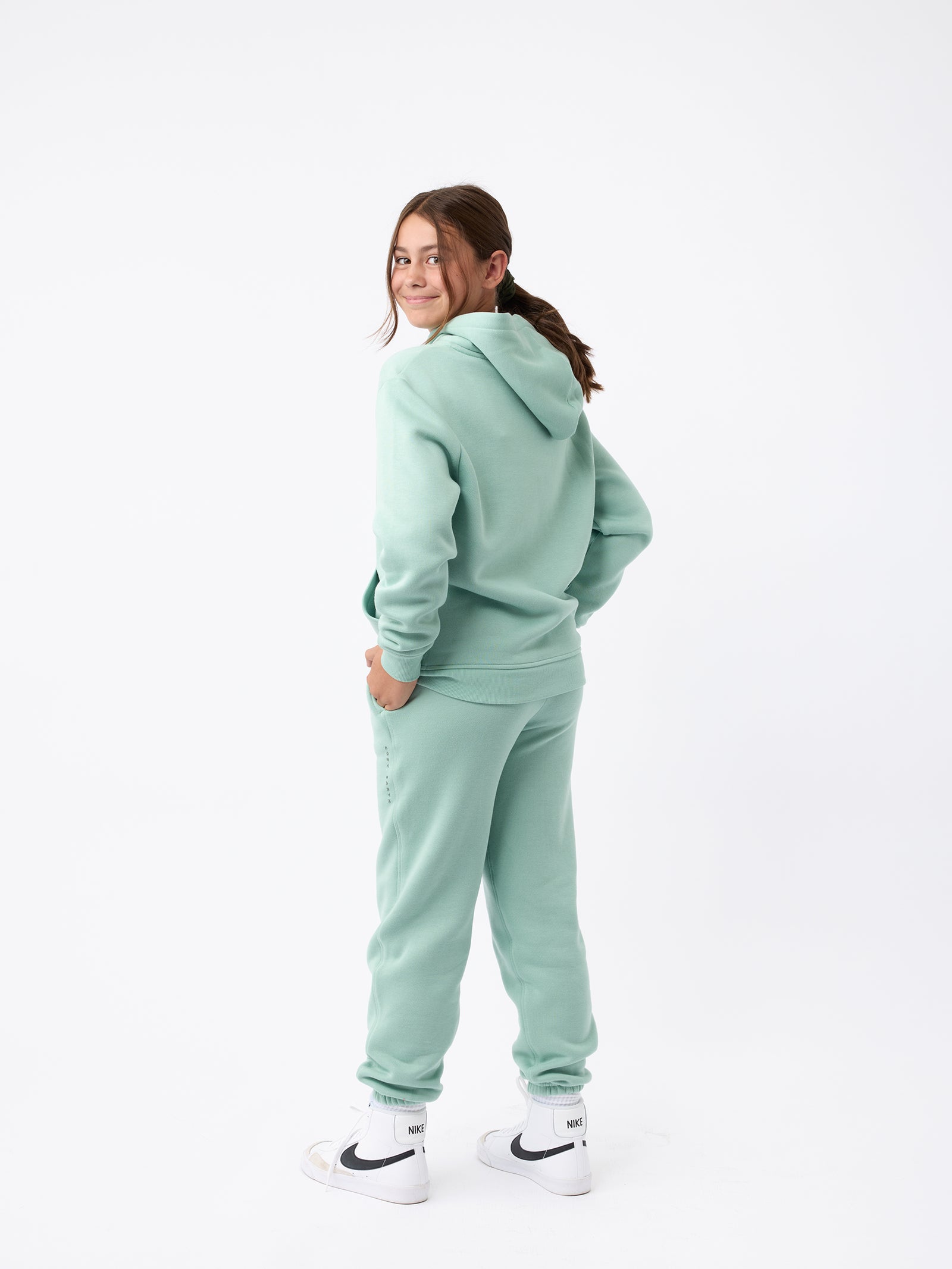 A person is wearing the Kid's CityScape Sweatpant in light green from Cozy Earth, paired with a matching hoodie. They stand in profile, facing right, with their hands tucked into their pockets. The outfit is completed with white high-top sneakers featuring black swooshes against a plain white background.