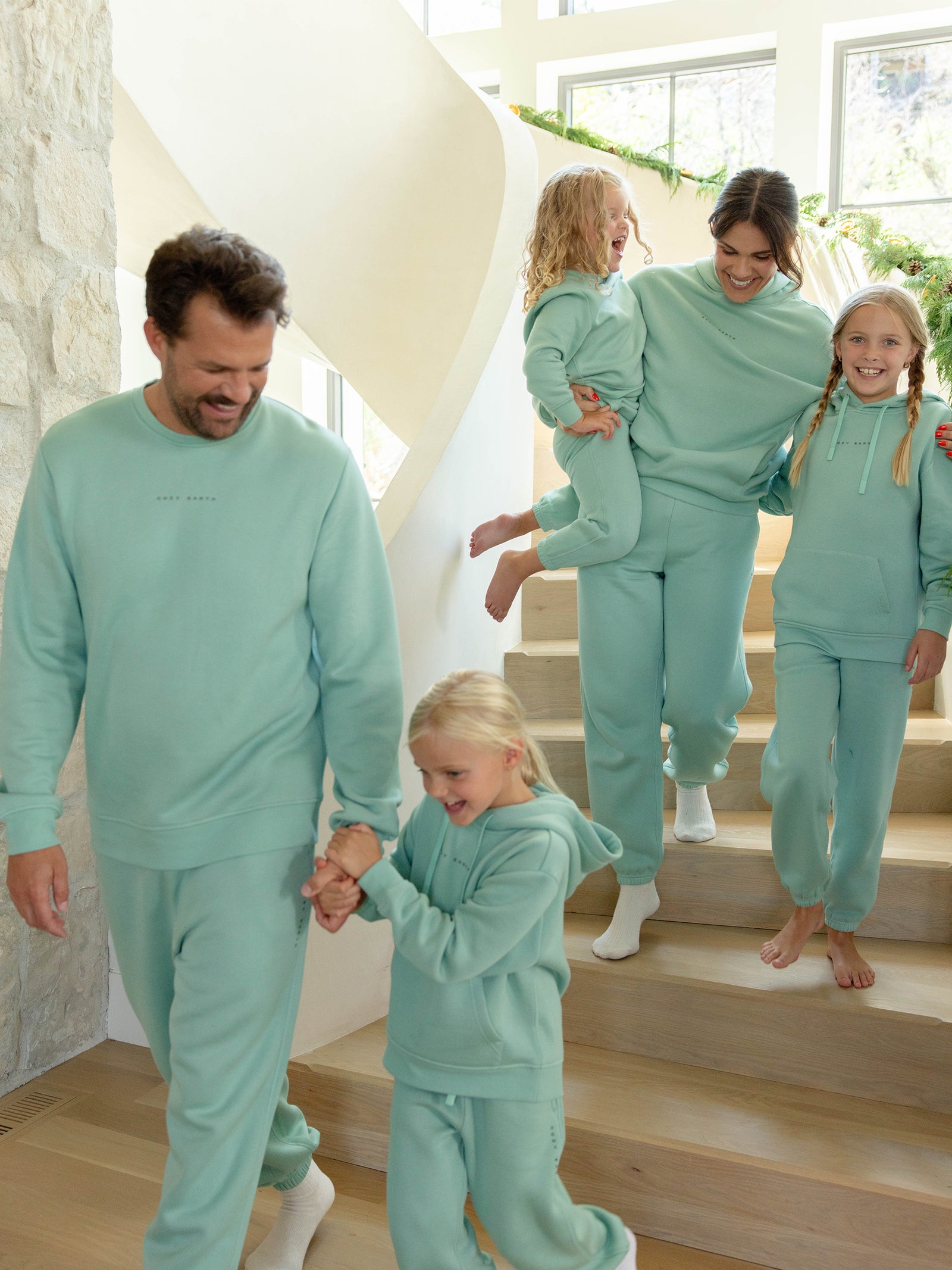 A family of five, clad in matching turquoise CityScape tracksuits by Cozy Earth, joyfully descends a modern staircase. The father, wearing the Men's CityScape Sweatpant, holds hands with one child at the front, while the mother carries another child and walks alongside their third child who is smiling brightly beside her.