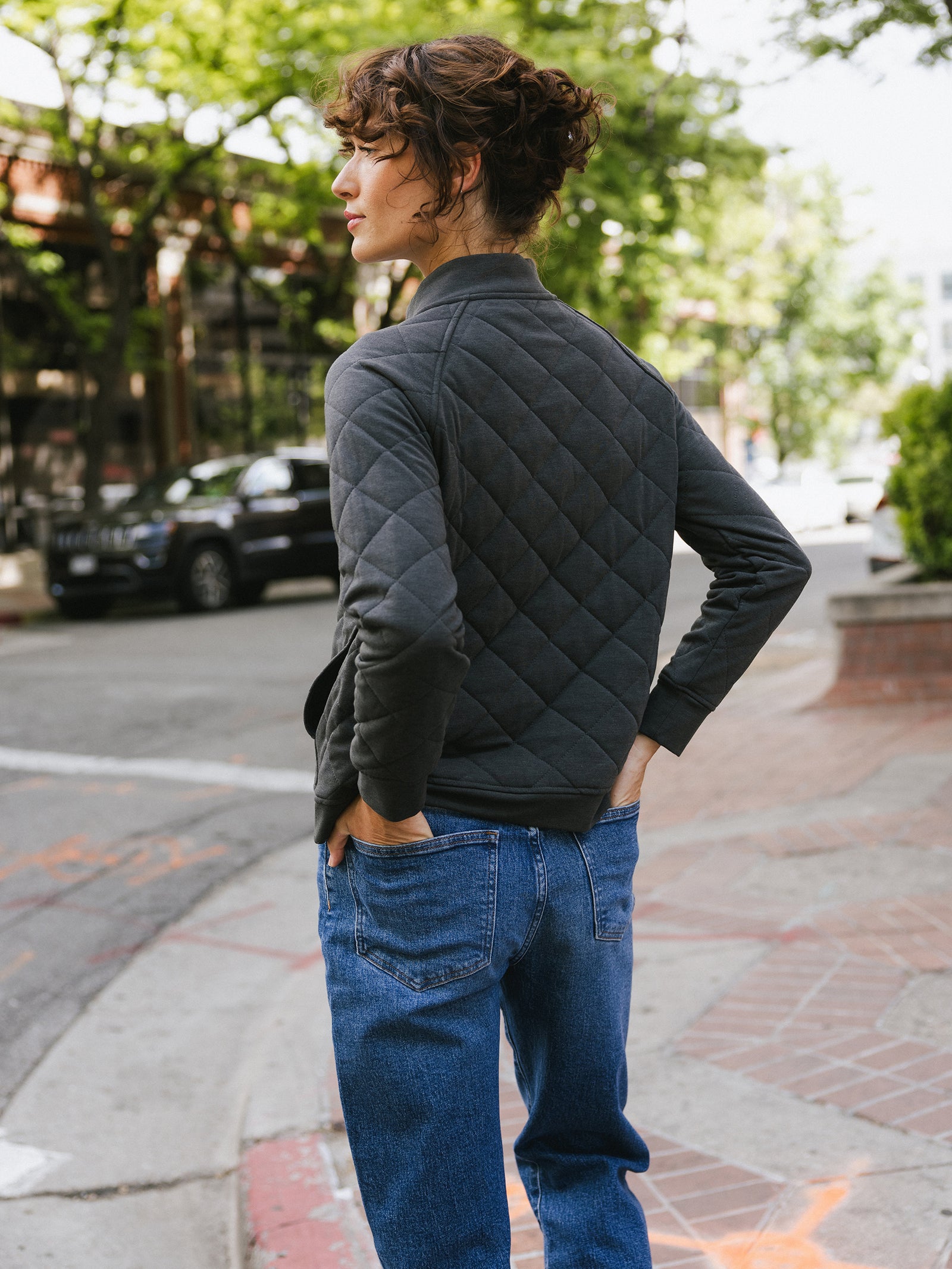 A person with curly hair stands on a city sidewalk, facing away from the camera. They are wearing a dark Women's Ultra-Soft Bamboo Quilted Snap Pullover by Cozy Earth and blue jeans. The background features trees, parked cars, and buildings. The scene appears to be in an urban area on a sunny day.