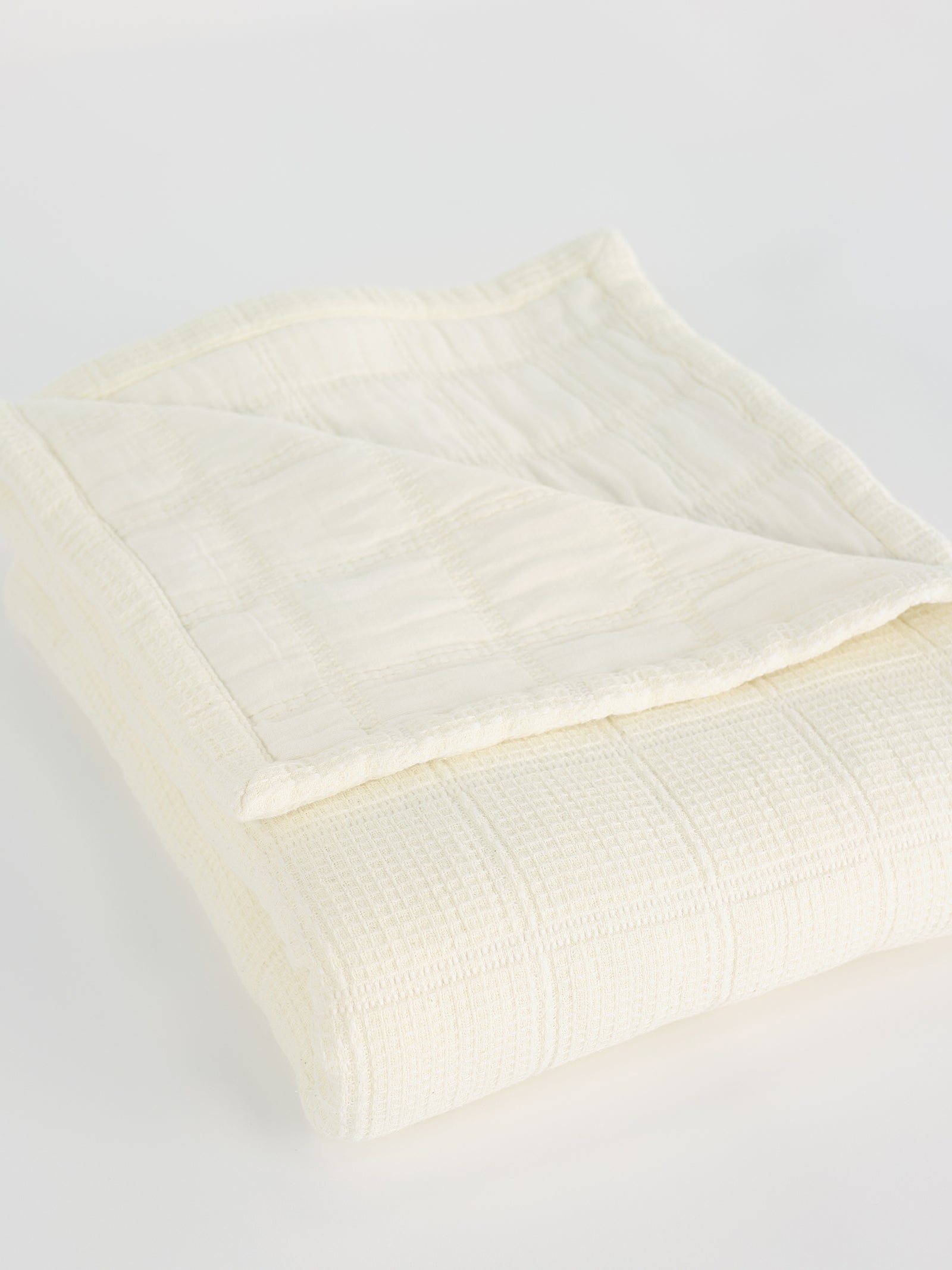 A neatly folded Waffle Windowpane Blanket from Cozy Earth, cream-colored with a subtle checkered pattern. The blanket has a soft texture and looks lightweight, perfect for adding a cozy layer to a bed or couch. The corner of the blanket is flipped over, revealing its delicate stitching and fabric detail.