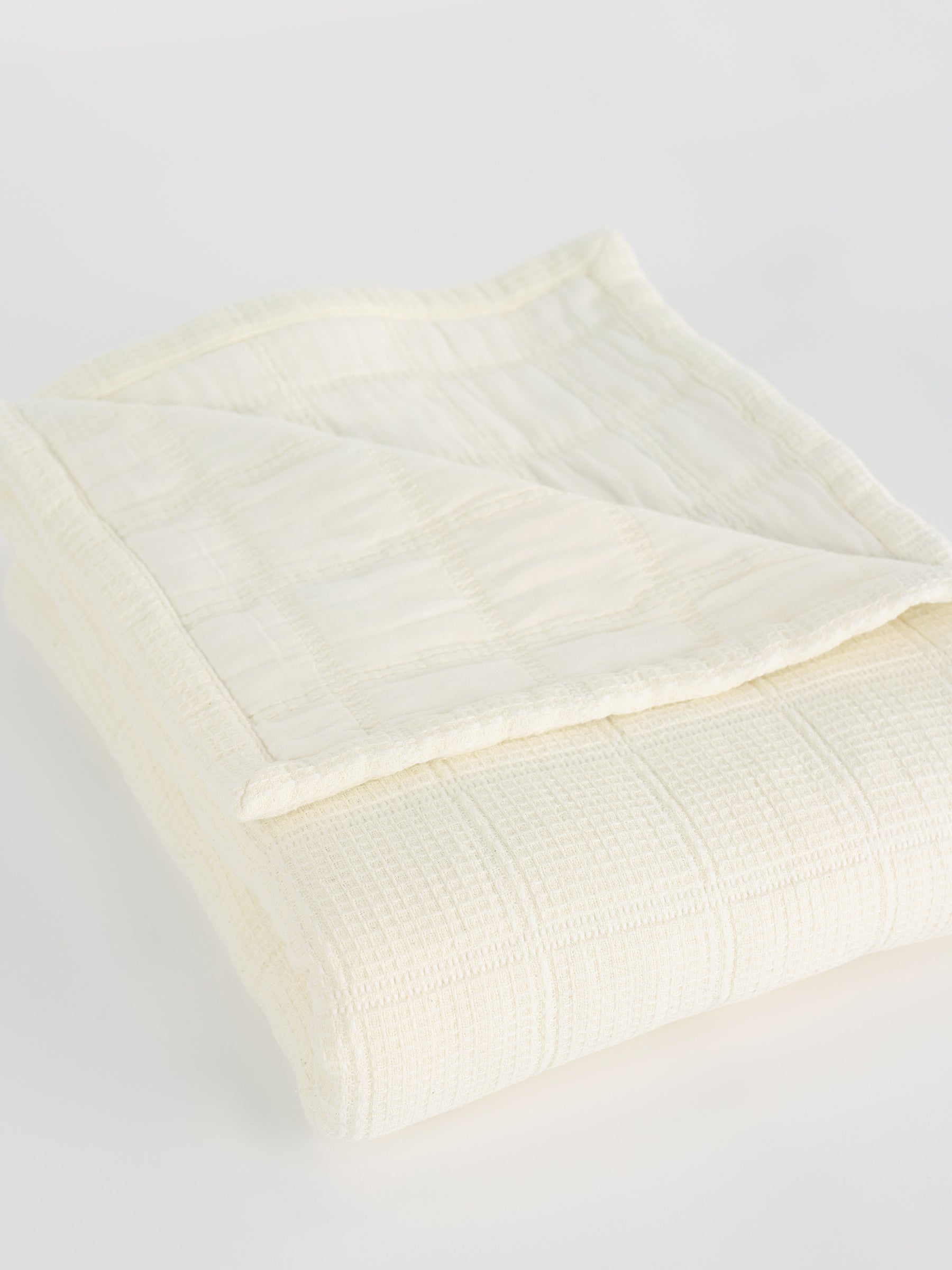A neatly folded Waffle Windowpane Blanket from Cozy Earth, cream-colored with a subtle checkered pattern. The blanket has a soft texture and looks lightweight, perfect for adding a cozy layer to a bed or couch. The corner of the blanket is flipped over, revealing its delicate stitching and fabric detail. |Color:Creme