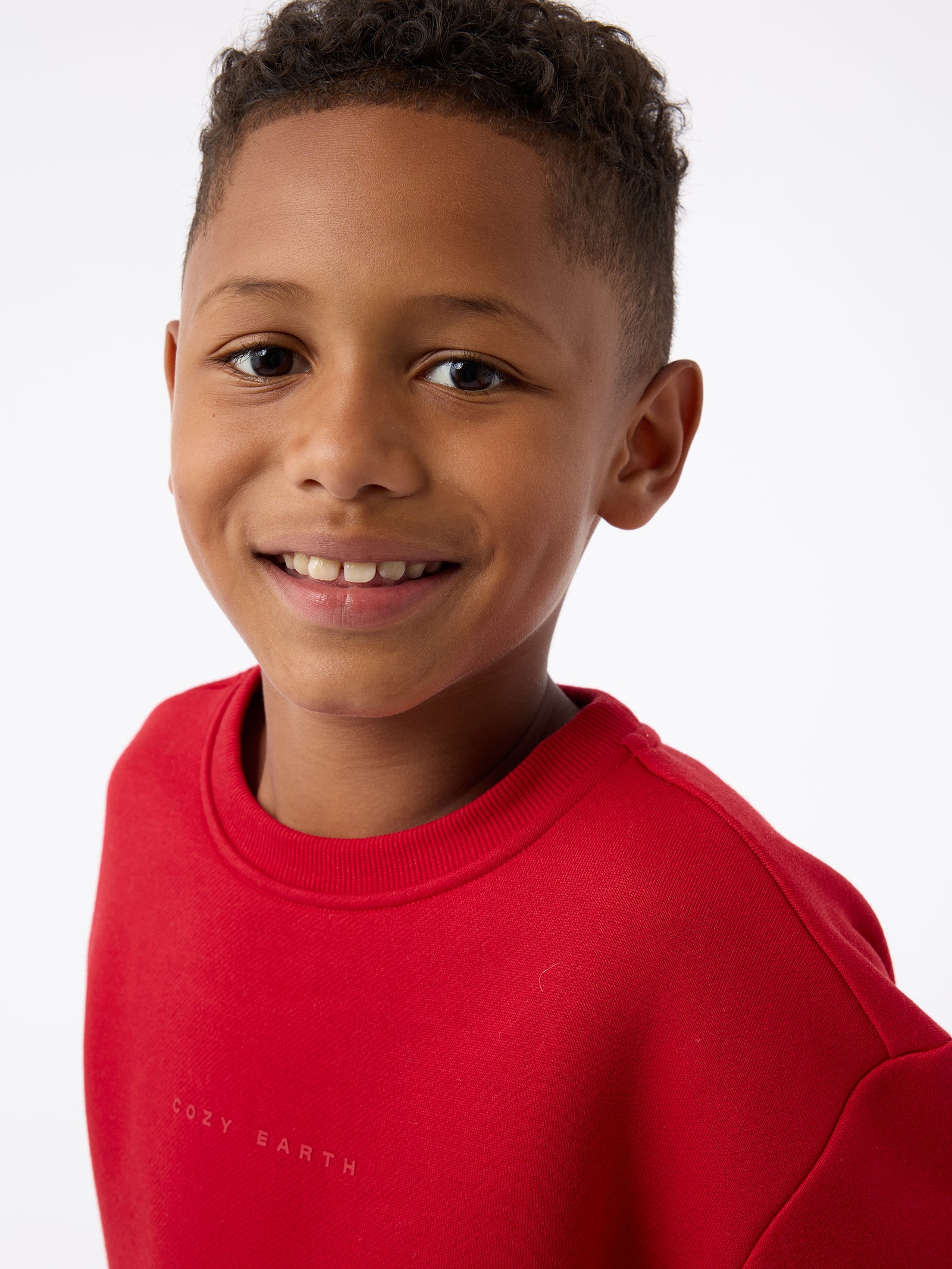 A young boy, dressed in a Cozy Earth Kid's CityScape Crewneck, smiles warmly at the camera against a white background.
