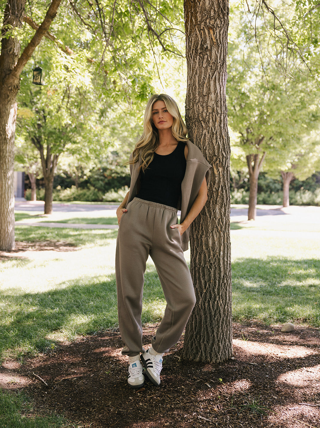 A person with long, wavy hair wearing a black tank top, Cozy Earth's Women's CityScape Sweatpant, and white sneakers stands against a tree trunk outdoors. The background features green grass, other trees, and sunlight filtering through the foliage.