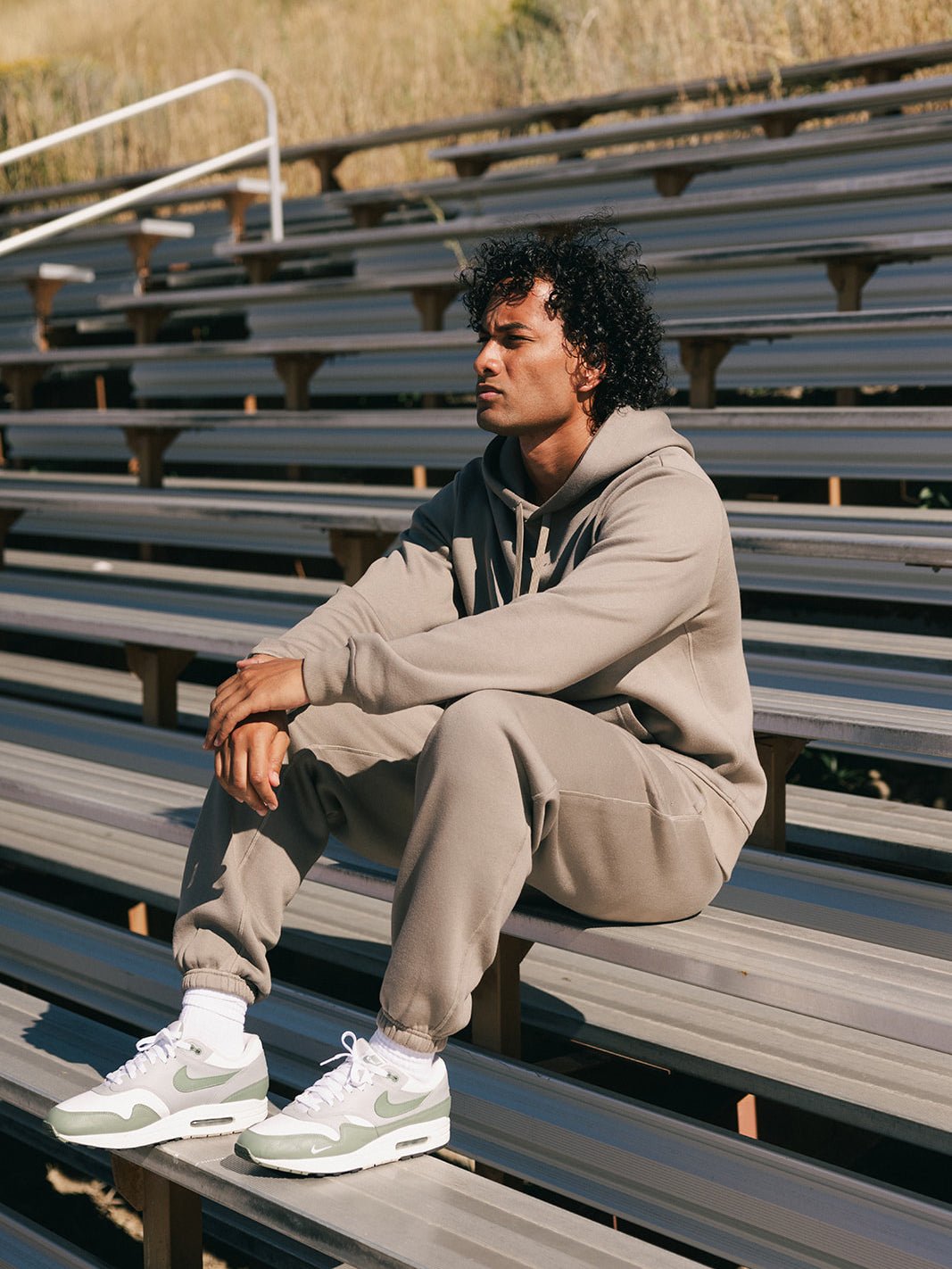 A person with curly hair sits on an empty set of bleachers outdoors, wearing a light gray hoodie and matching Cozy Earth Men's CityScape Sweatpants, paired with green and white Nike sneakers. They look off into the distance with a thoughtful expression.