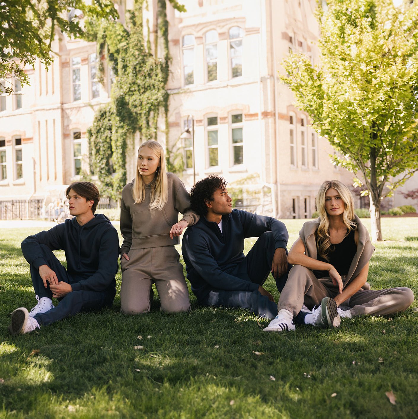 Four people, two men and two women, are sitting on grass in front of a building with large windows and walls covered in ivy. They are dressed in casual athletic wear, with the men sporting the Men's CityScape Quarter Zip from Cozy Earth. The scene suggests a relaxed and friendly atmosphere on a sunny day.
