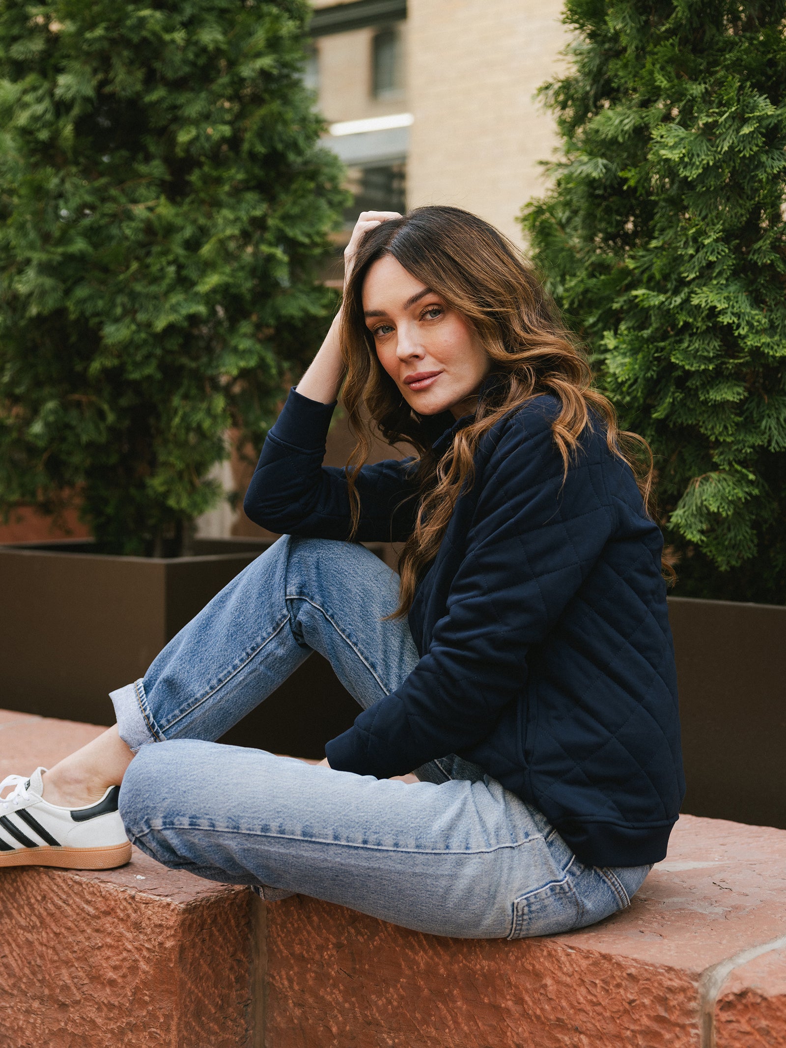 A woman with long, wavy brown hair sits casually on a red stone ledge. She is wearing a Cozy Earth Women's Ultra-Soft Bamboo Quilted Snap Pullover, light blue jeans, and white sneakers with black stripes. She rests her chin on her hand and looks at the camera, with green foliage in the background.