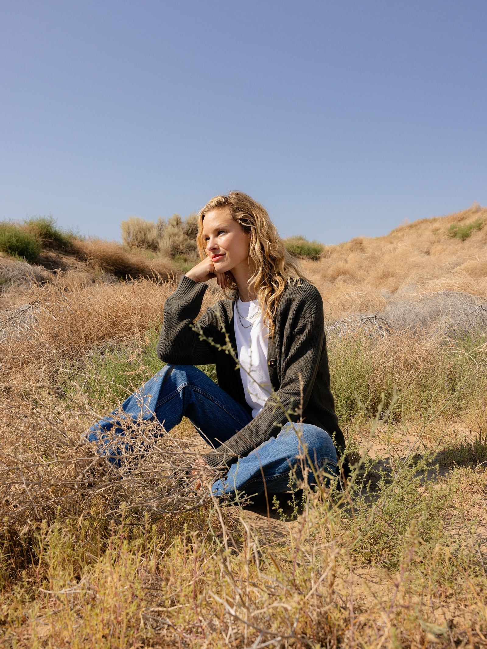 A person with curly hair, wearing a white shirt, Cozy Earth's Oversized Classic Cardigan in green, and jeans, is sitting thoughtfully on a grassy hillside under a clear blue sky. The scene is serene, with dry grass and sparse bushes in the background.