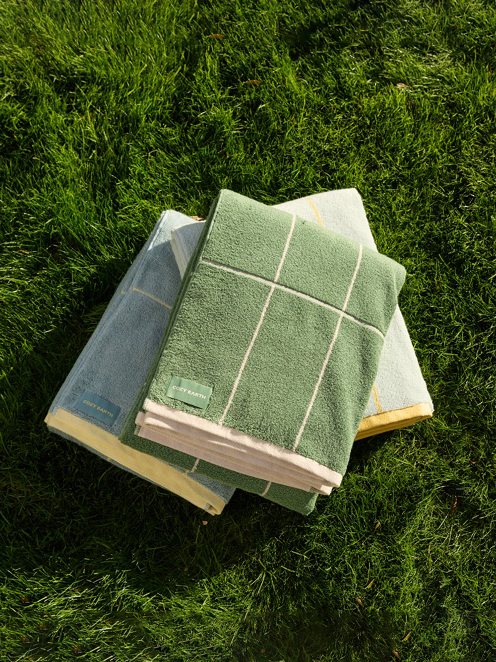 Three folded Cozy Earth Windowpane Resort Towels, in green, blue, and light yellow with small green labels attached, rest on lush grass. Sunlight creates shadows that accentuate the texture of both the towels and the grass.