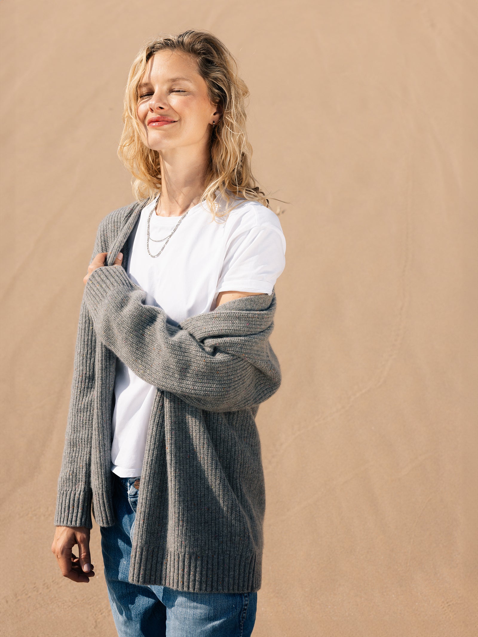 A person stands in a sandy desert, wearing a white T-shirt, an Oversized Sunday Cashmere Cardigan from Cozy Earth, and jeans. They have long, wavy blonde hair and a relaxed expression, with eyes closed and a slight smile. The sun casts a soft light on them.