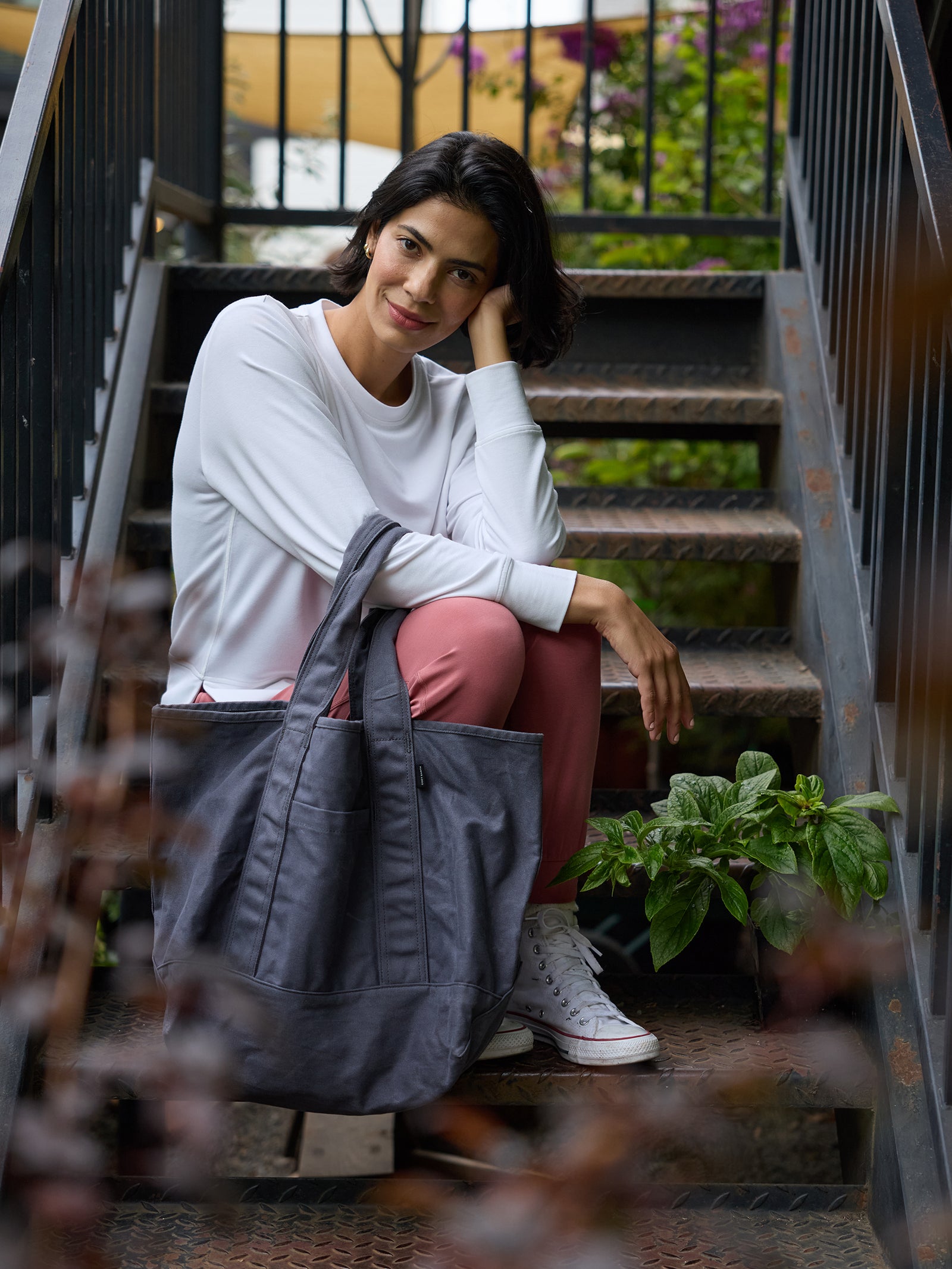 A woman with short, dark hair sits on metal stairs outdoors, resting her head on her hand. She is wearing a white sweater, pink pants, and white sneakers, and is holding a large Waxed Canvas Tote from Cozy Earth. Green foliage is visible in the background.
