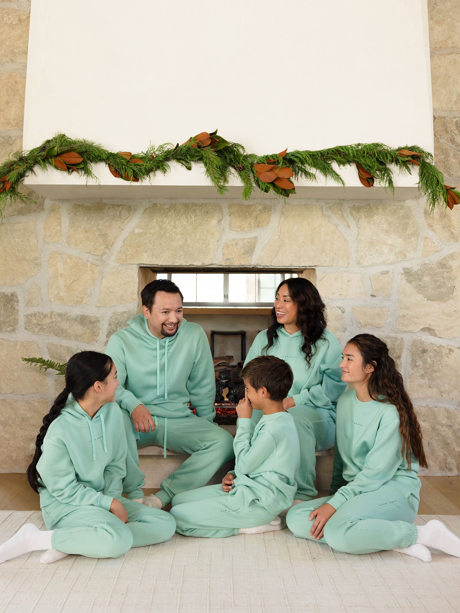 A family of five, consisting of two adults and three children, sits in front of a stone fireplace adorned with greenery. The children are wearing matching mint green Kid's CityScape Sweatpants by Cozy Earth. They are all smiling at each other, creating a warm and joyful atmosphere.
