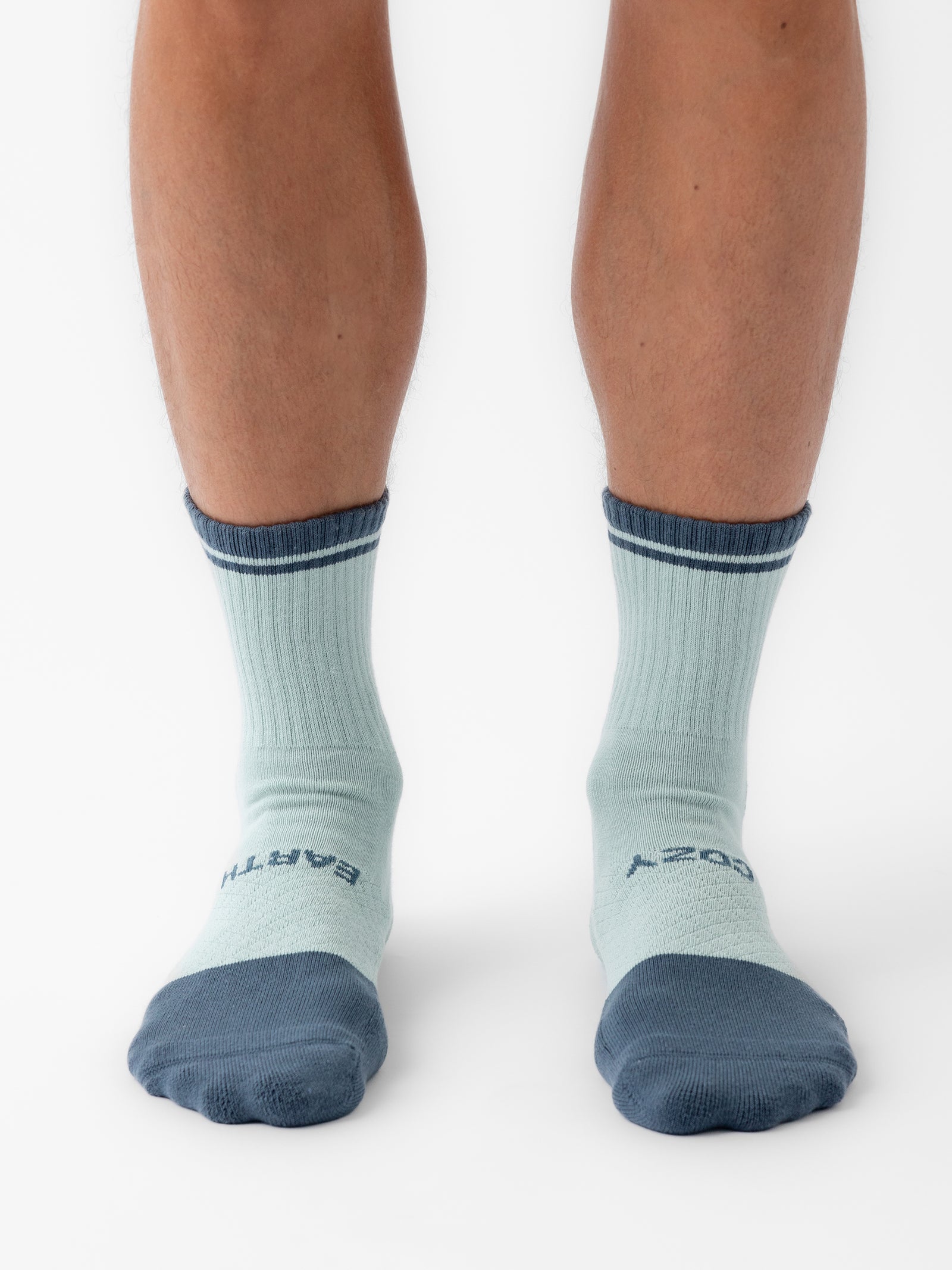 A person wearing light blue Essential Quarter Socks from Cozy Earth, featuring a dark blue toe, heel, and cuff, stands on a white background. The socks have "BARTER" and "COZY" written on each foot, with the person's lower legs and feet visible.